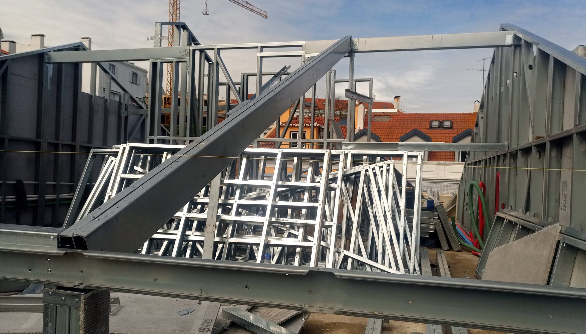 Canteiro de obras com estrutura de aço. Vigas de sustentação cinza, estruturas de aço sobrepostas nas paredes e telhado vermelho ao fundo.