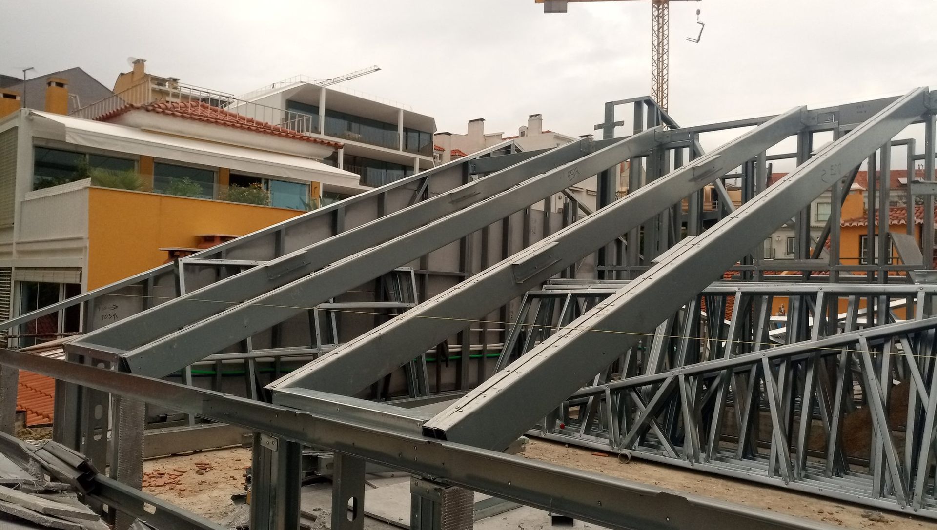 Construção de telhado com estrutura metálica em um canteiro de obras, com outros edifícios e um guindaste visíveis ao fundo.