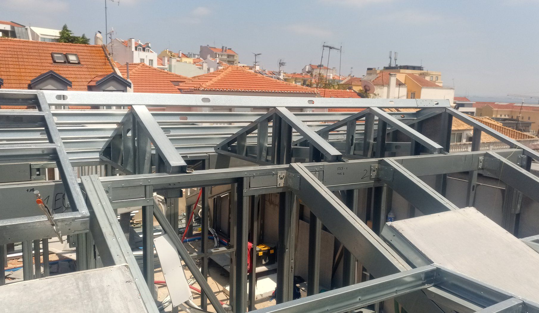 Estrutura metálica de um edifício em construção, com telhados e a linha do horizonte da cidade ao fundo.