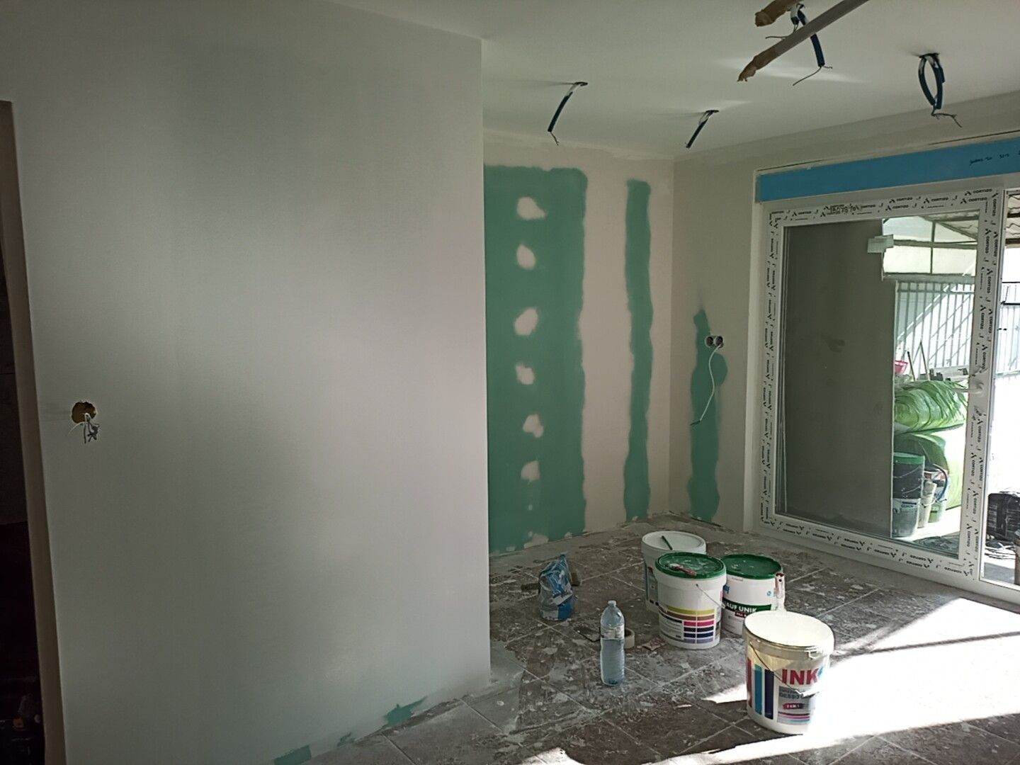 Quarto em reforma com drywall e latas de tinta; janela ao fundo.