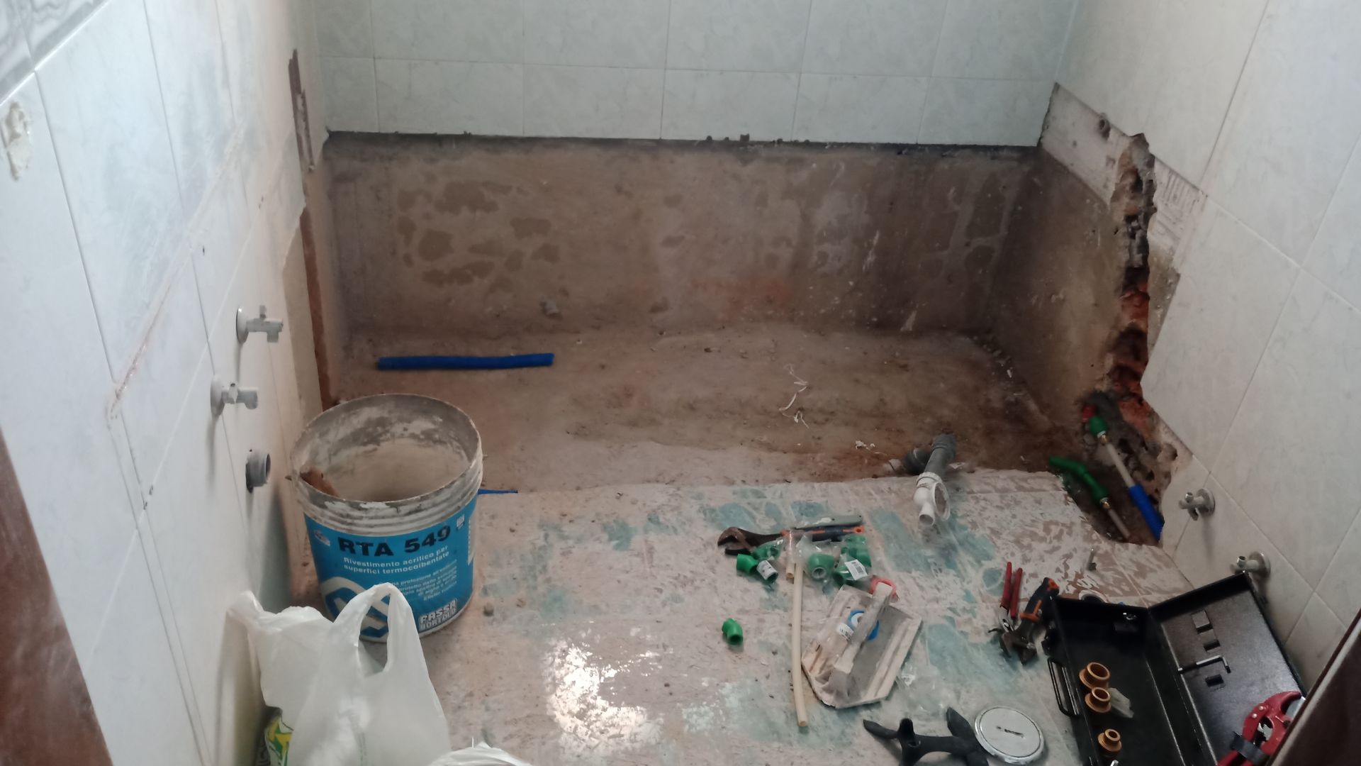 Banheiro em construção: paredes com azulejos, piso de cimento, ferramentas, balde de tinta.