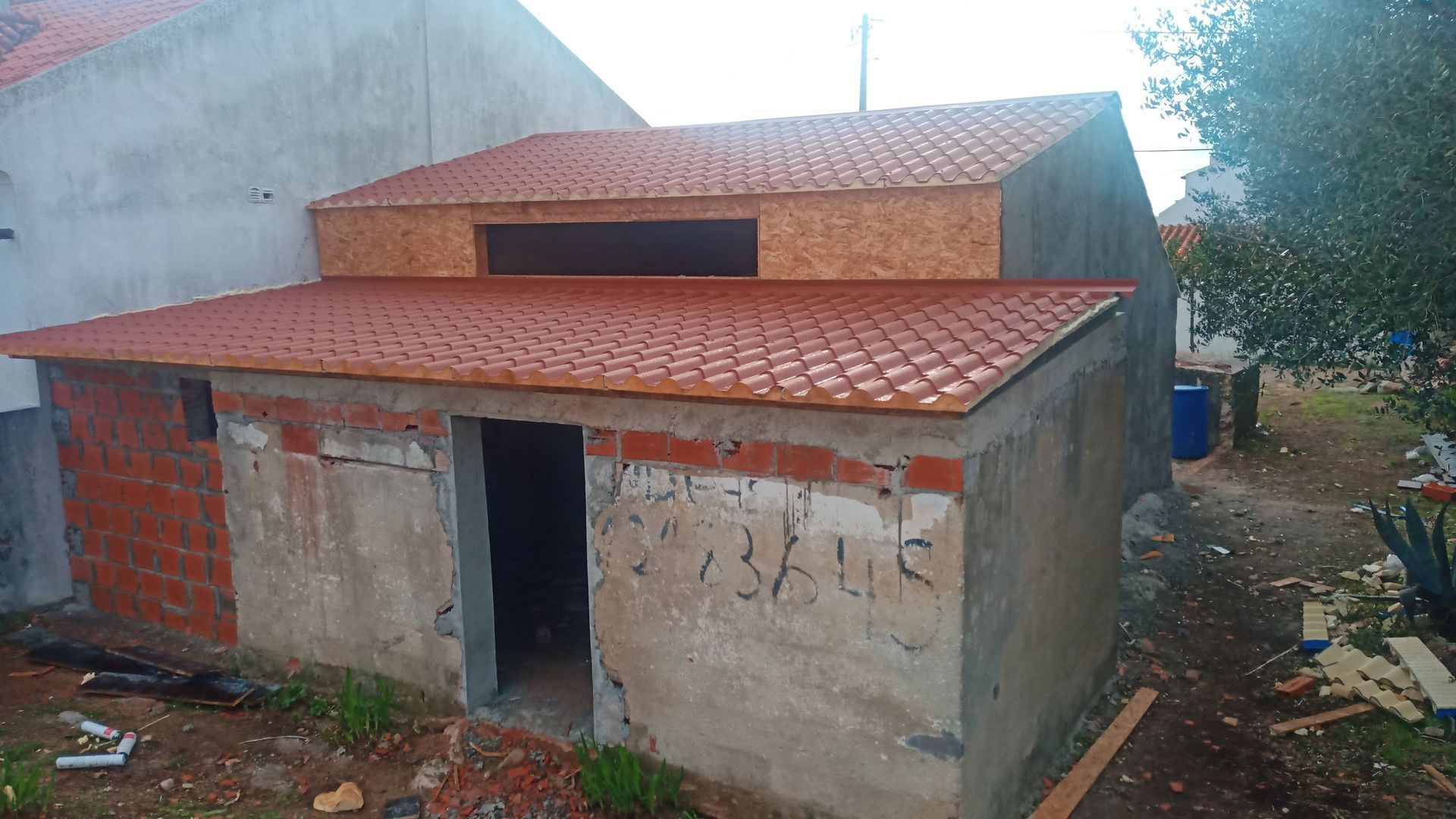 Pequeno edifício inacabado de concreto e tijolo com telhado de telha vermelha, ambiente externo.