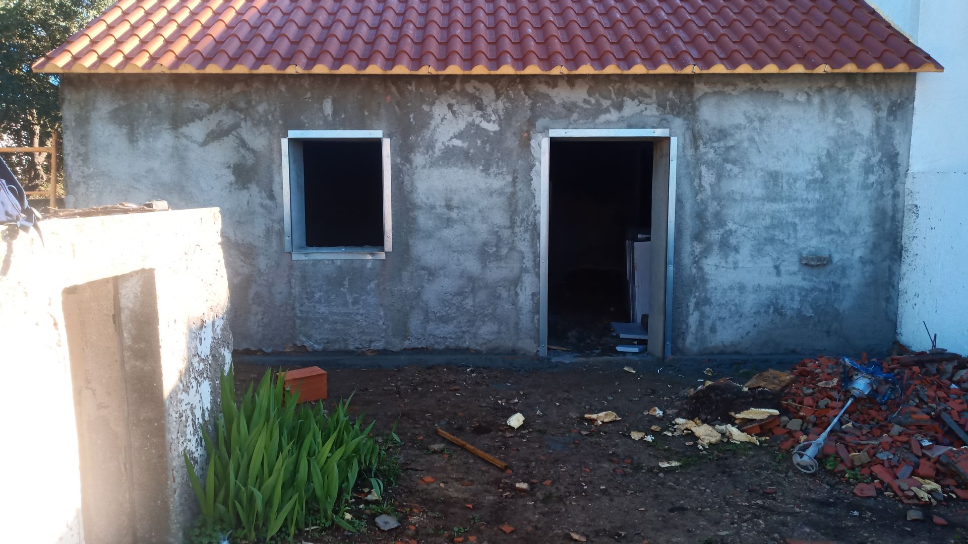 Pequena construção inacabada com telhado de telhas vermelhas, janela e porta. Materiais de construção e entulho no chão.