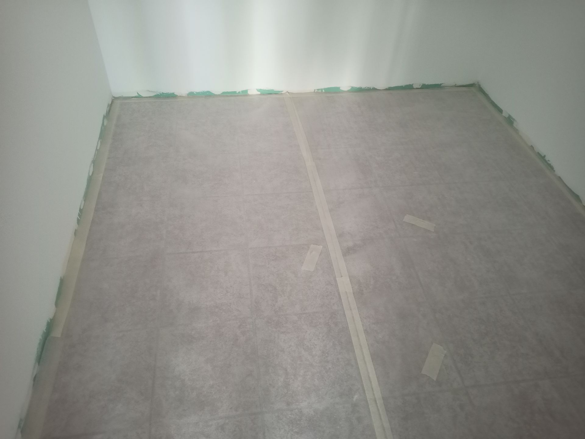 Quarto com o chão coberto de papel cinza e fita verde nas bordas. As paredes são brancas.