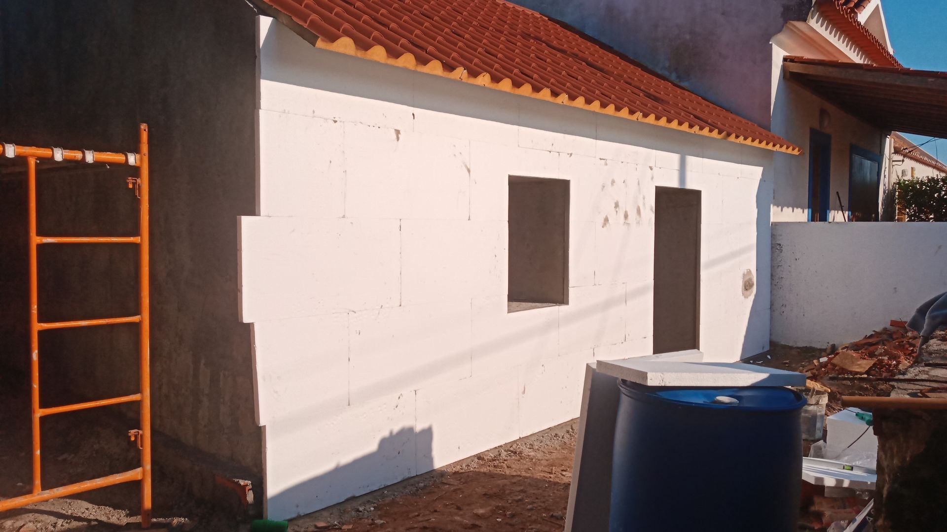 Exterior de um edifício em construção, paredes brancas, telhado vermelho, andaimes laranja, duas janelas.