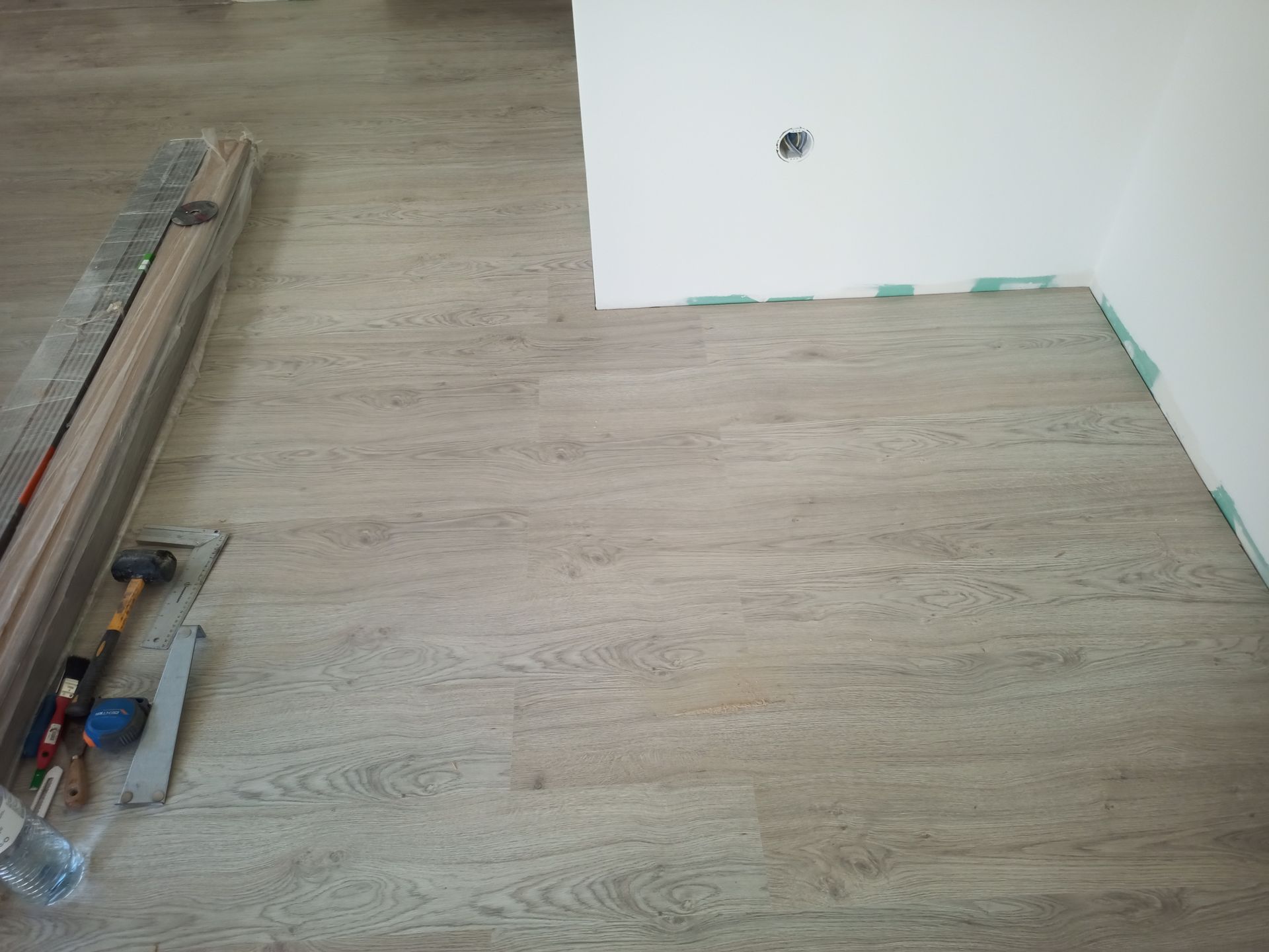 Piso laminado claro com aparência de madeira instalado em um cômodo, com ferramentas e tábuas ainda fechadas por perto. Parede branca com tomada elétrica.