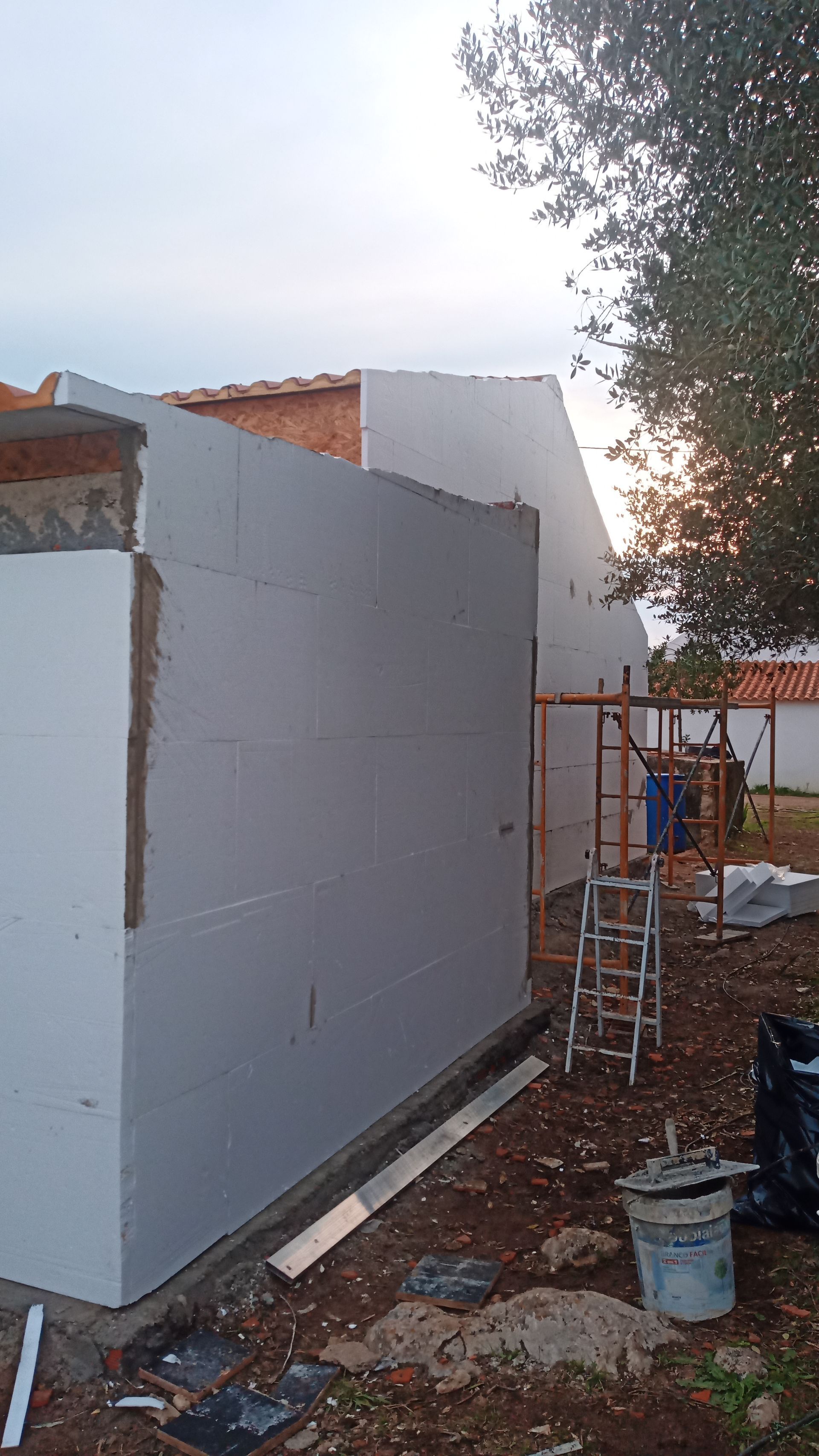 Exterior de um prédio com isolamento de espuma branca, andaimes e um operário. Céu nublado.