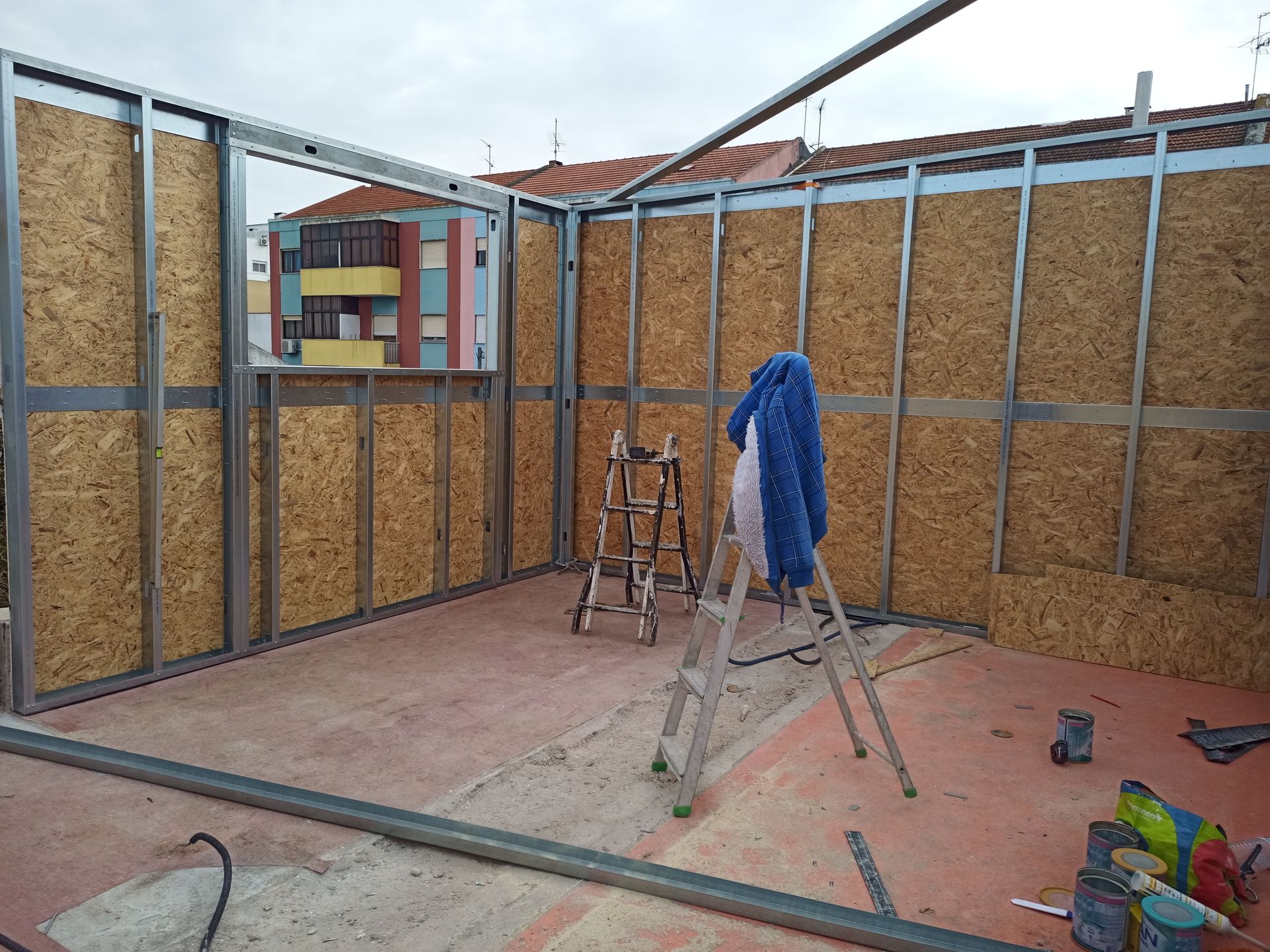 Construção interna: paredes com estrutura metálica e placas de OSB. Piso de concreto vermelho. Escadas e ferramentas disponíveis.