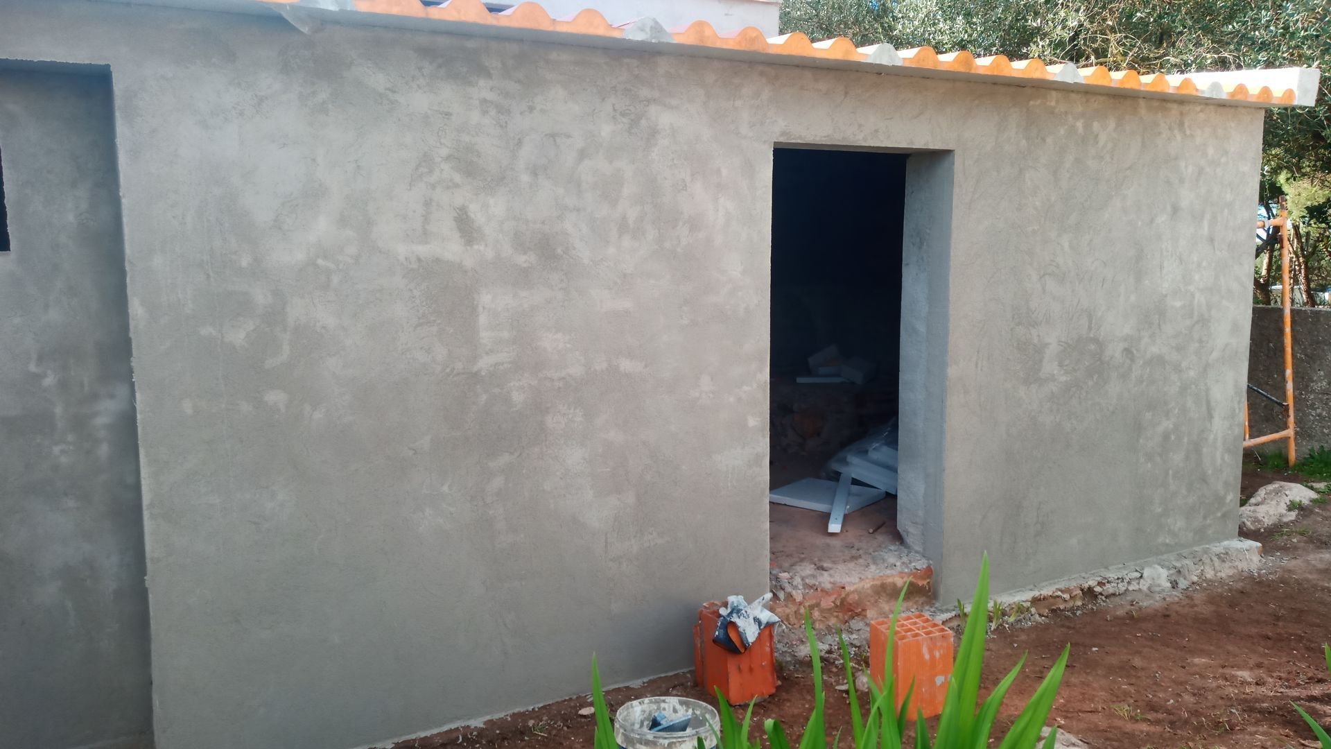 Pequeno prédio cinza de estuque com porta aberta e telhado de zinco; foto do exterior.
