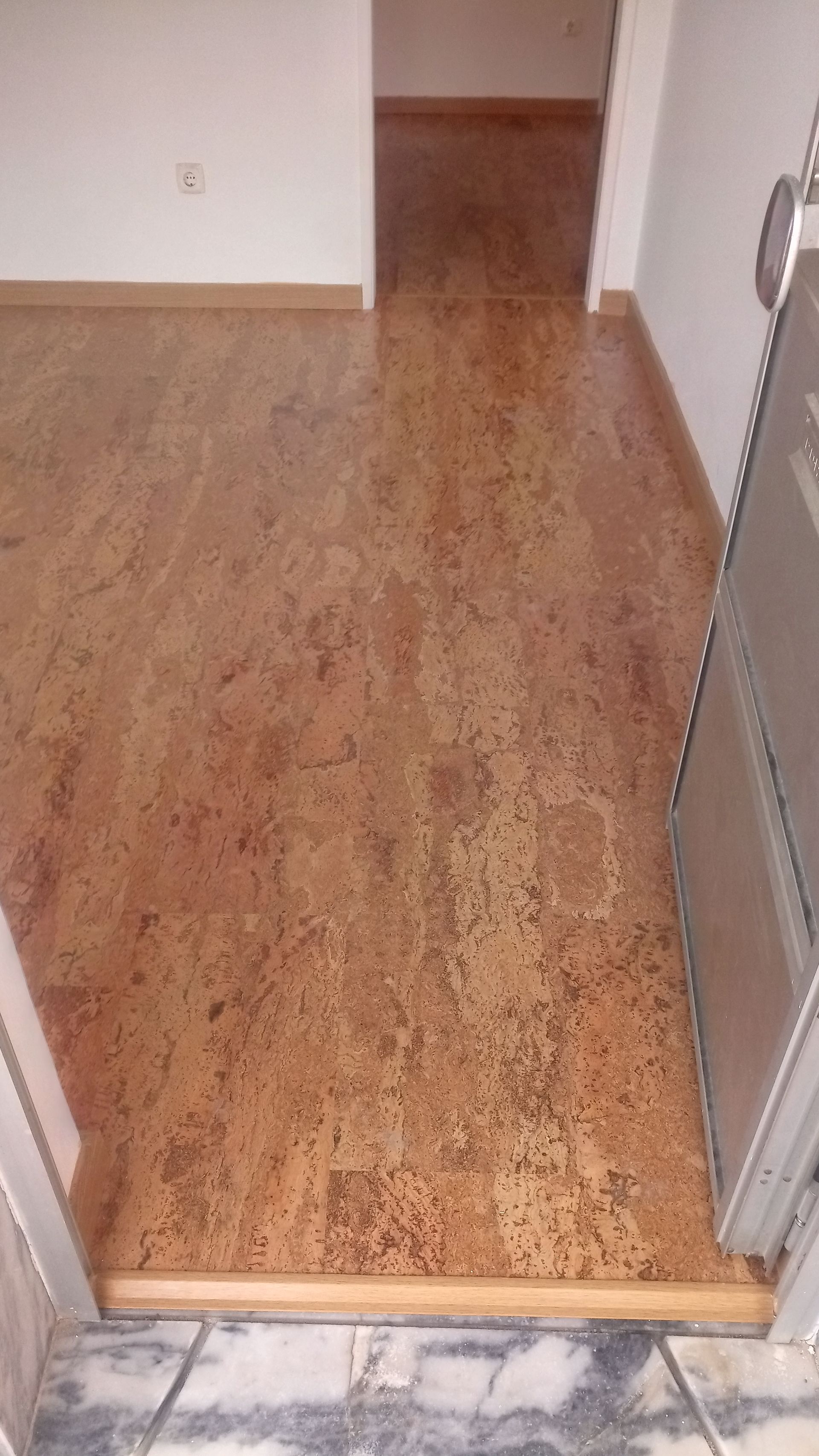 Piso de cortiça marrom em um corredor, com porta e vão de entrada visíveis.