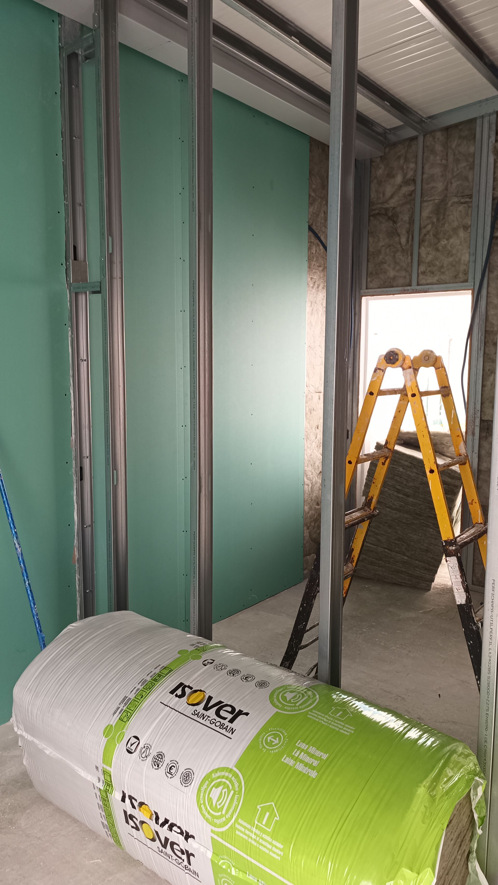 Canteiro de obras interno com estrutura metálica, drywall verde, rolo de isolamento e escada.