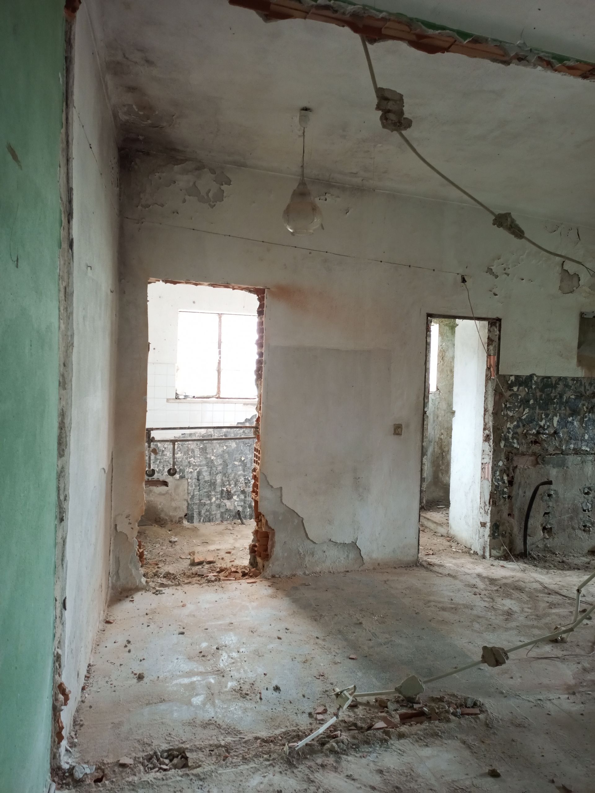 Interior de um quarto dilapidado com tijolos e portas expostas; luminária pendurada no teto.