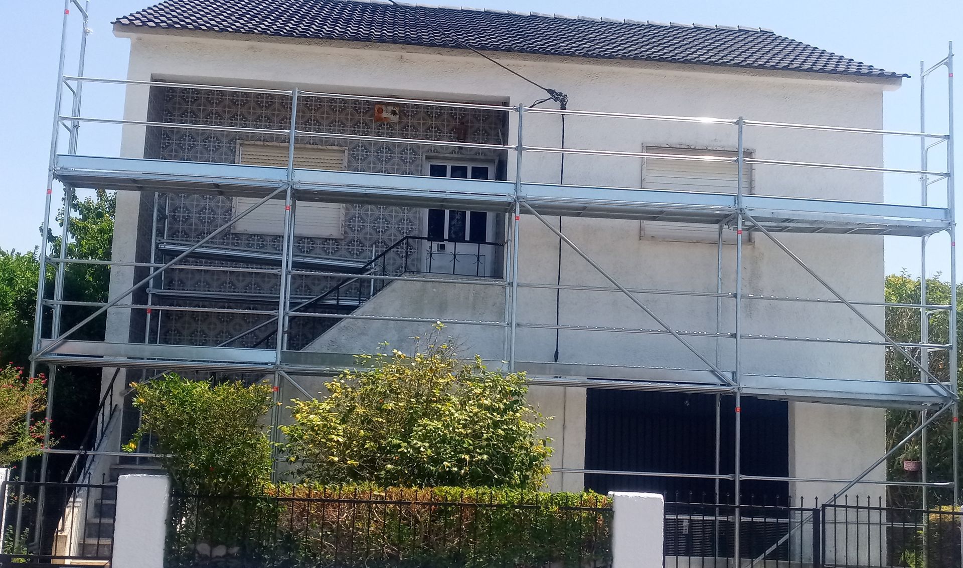 Andaimes em volta de uma casa de dois andares em reforma. A casa tem fachada de pedra e venezianas pretas.