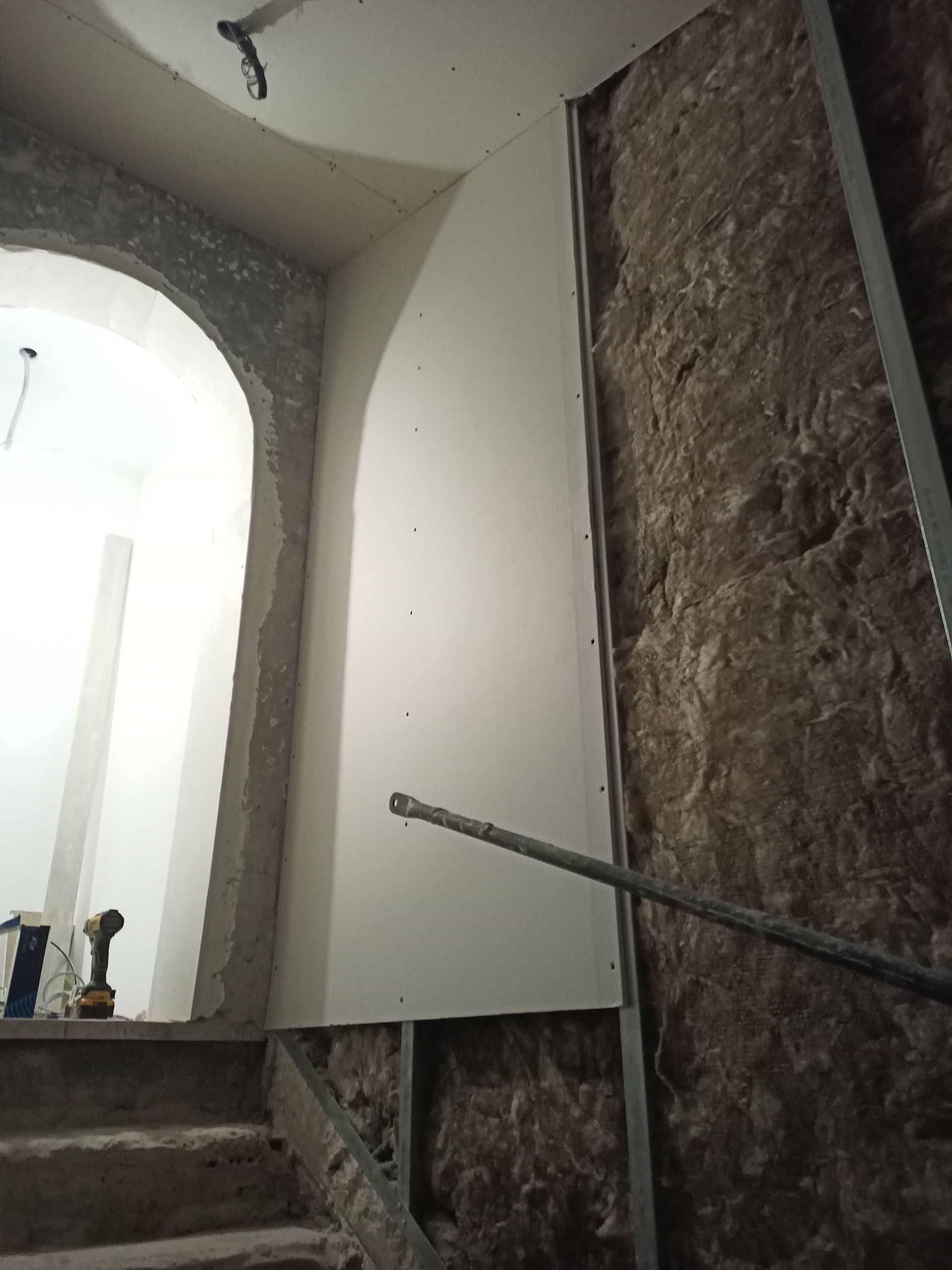 Construção interna com drywall em uma parede arqueada. Perfis metálicos, isolamento e escadas são visíveis.
