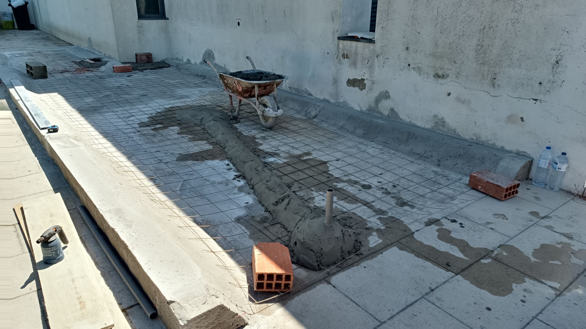 Canteiro de obras com concreto fresco em um espaço retangular, com carrinho de mão, tijolos e ferramentas presentes.