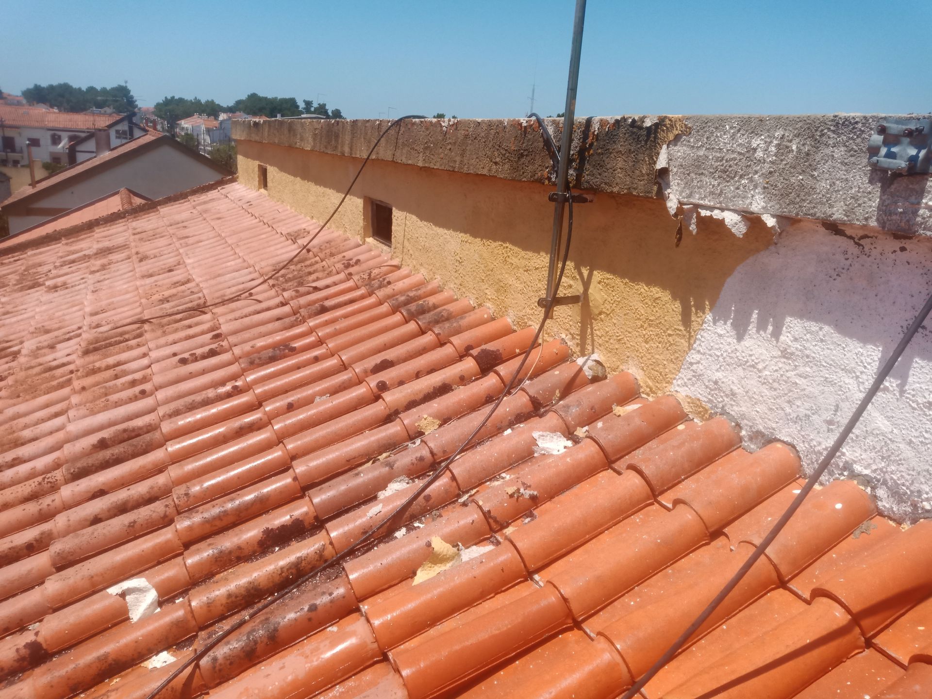 Telhado de telhas vermelhas com beiral danificado, cabos e vista parcial da cidade ao fundo.