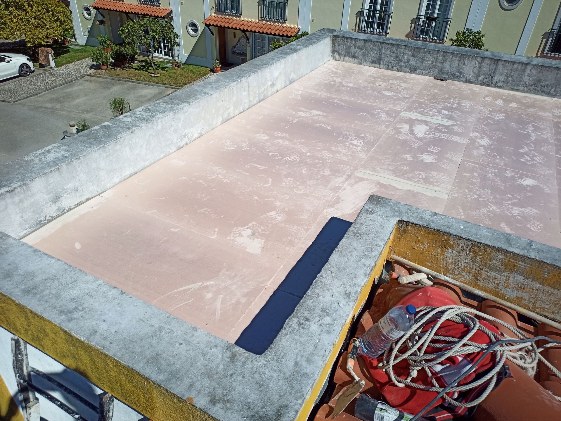 Telhado plano, de cor rosa-claro, com bordas de concreto. Alguns equipamentos estão encostados em um canto; um prédio e um carro são visíveis ao fundo.