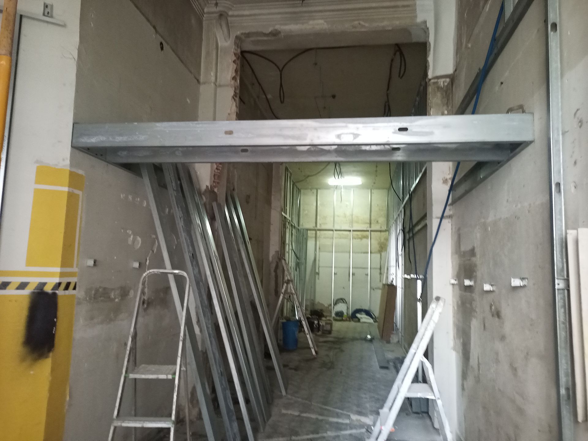 Obras em um corredor: vigas e perfis metálicos, escadas, fiação exposta e paredes inacabadas.