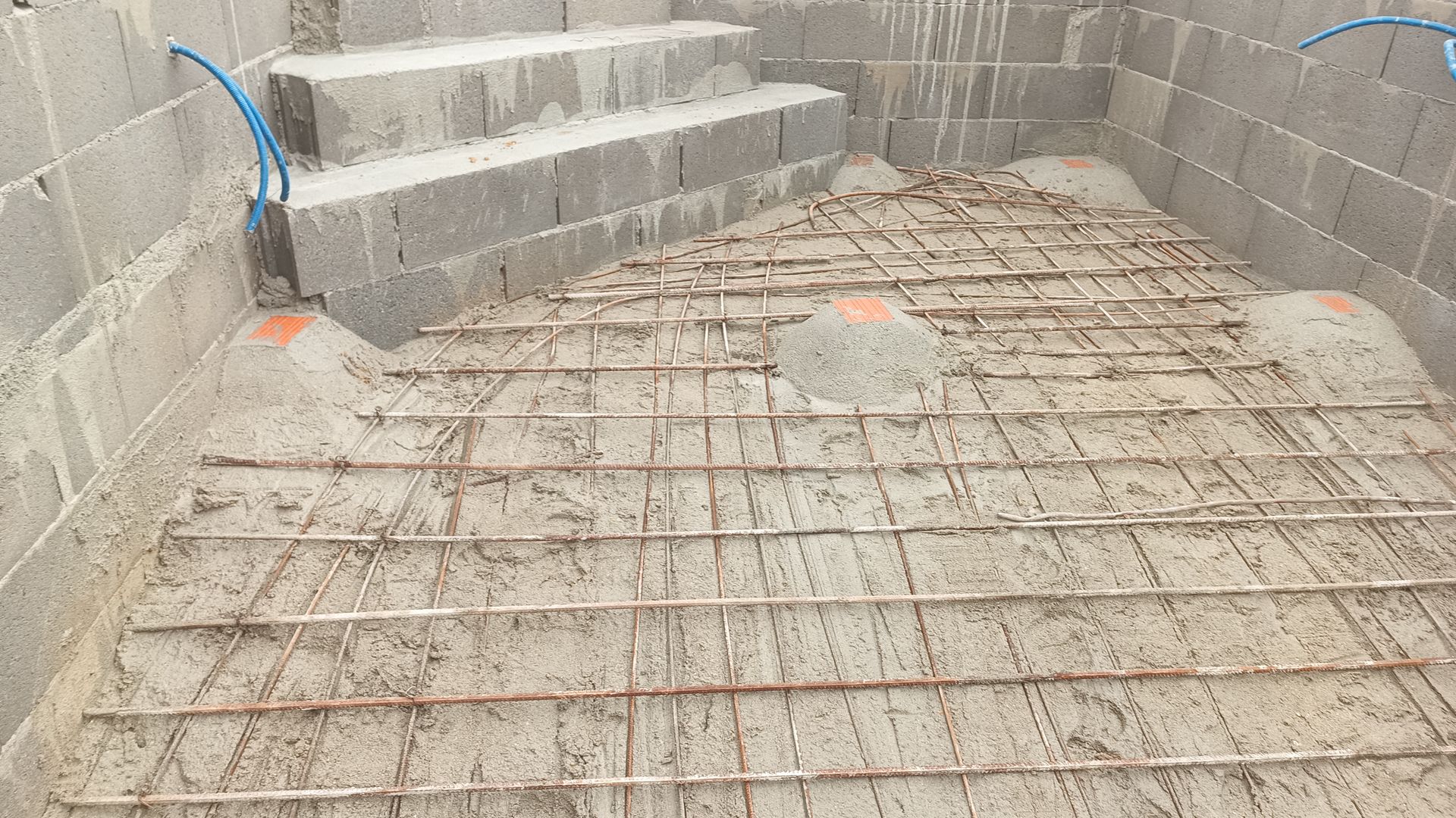Construção do interior da piscina com estrutura de vergalhões e degraus, paredes de blocos de concreto cinza.