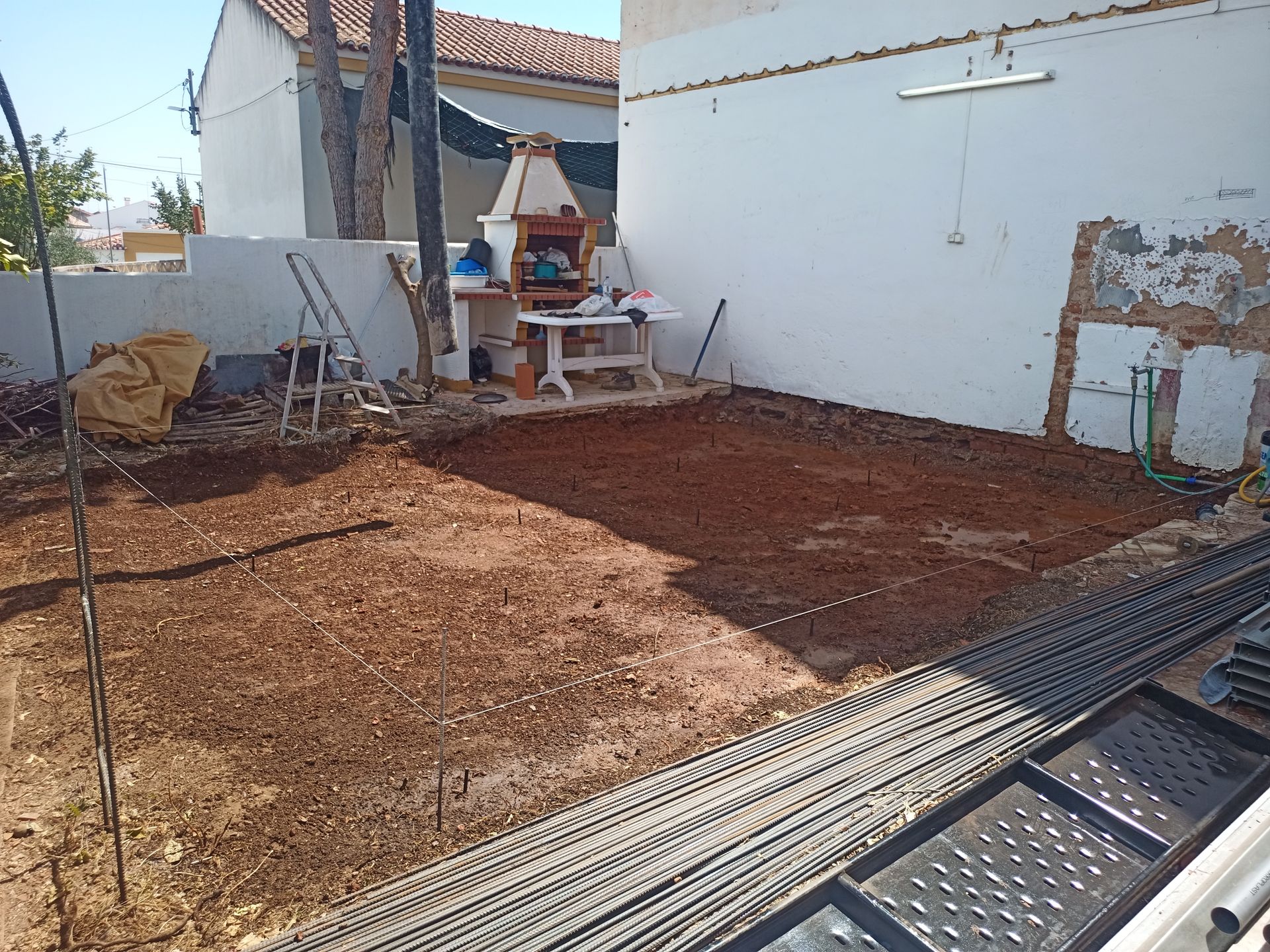 Canteiro de obras no quintal; terra nua com vergalhões e área para churrasqueira.