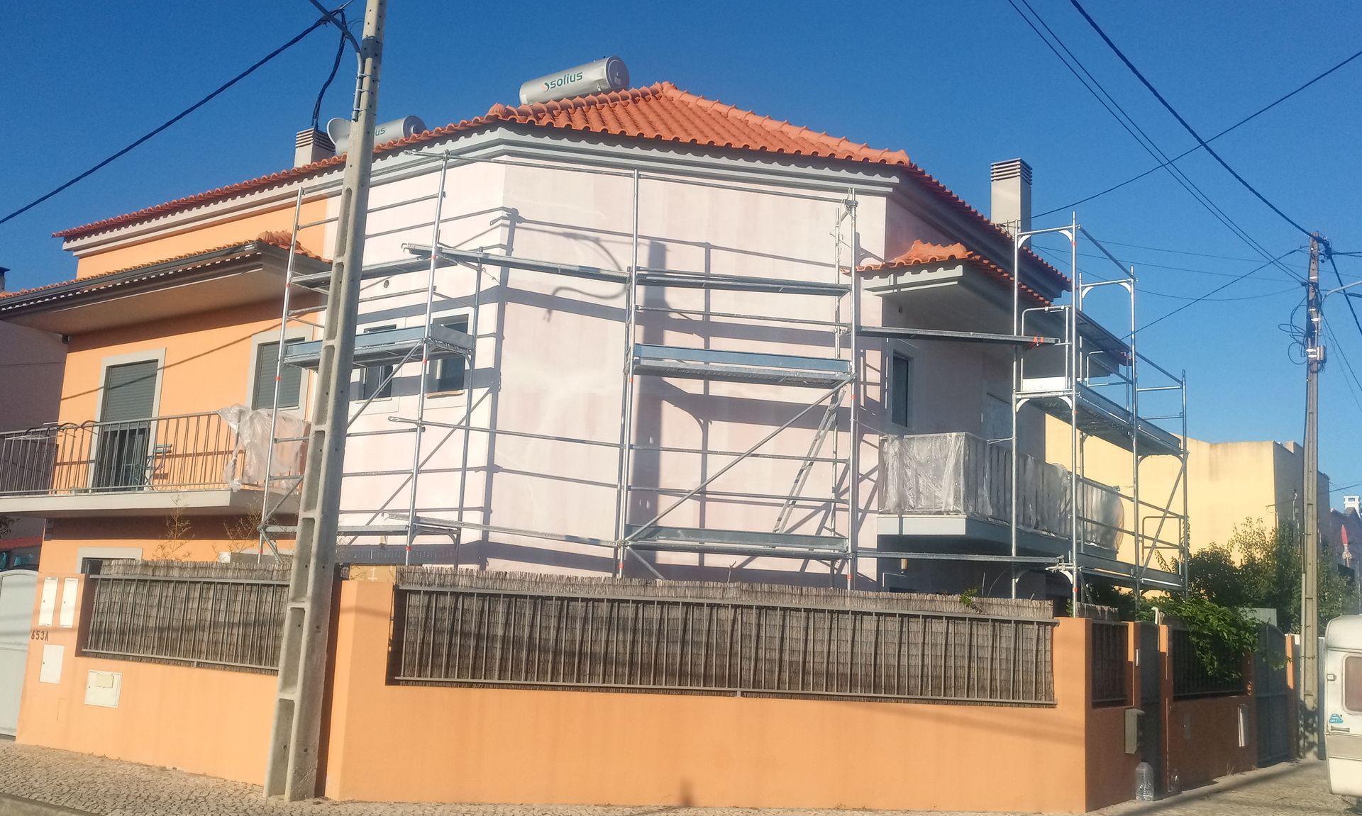 Exterior de uma casa com andaimes durante a reforma. Paredes rosa e laranja, telhado de telha vermelha, cerca de madeira.