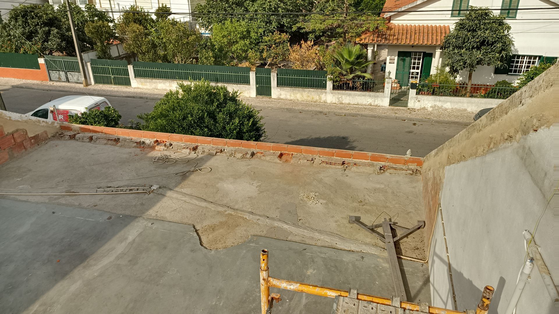 Vista do telhado de uma rua com casas e árvores. Um carro branco e andaimes de construção estão presentes.