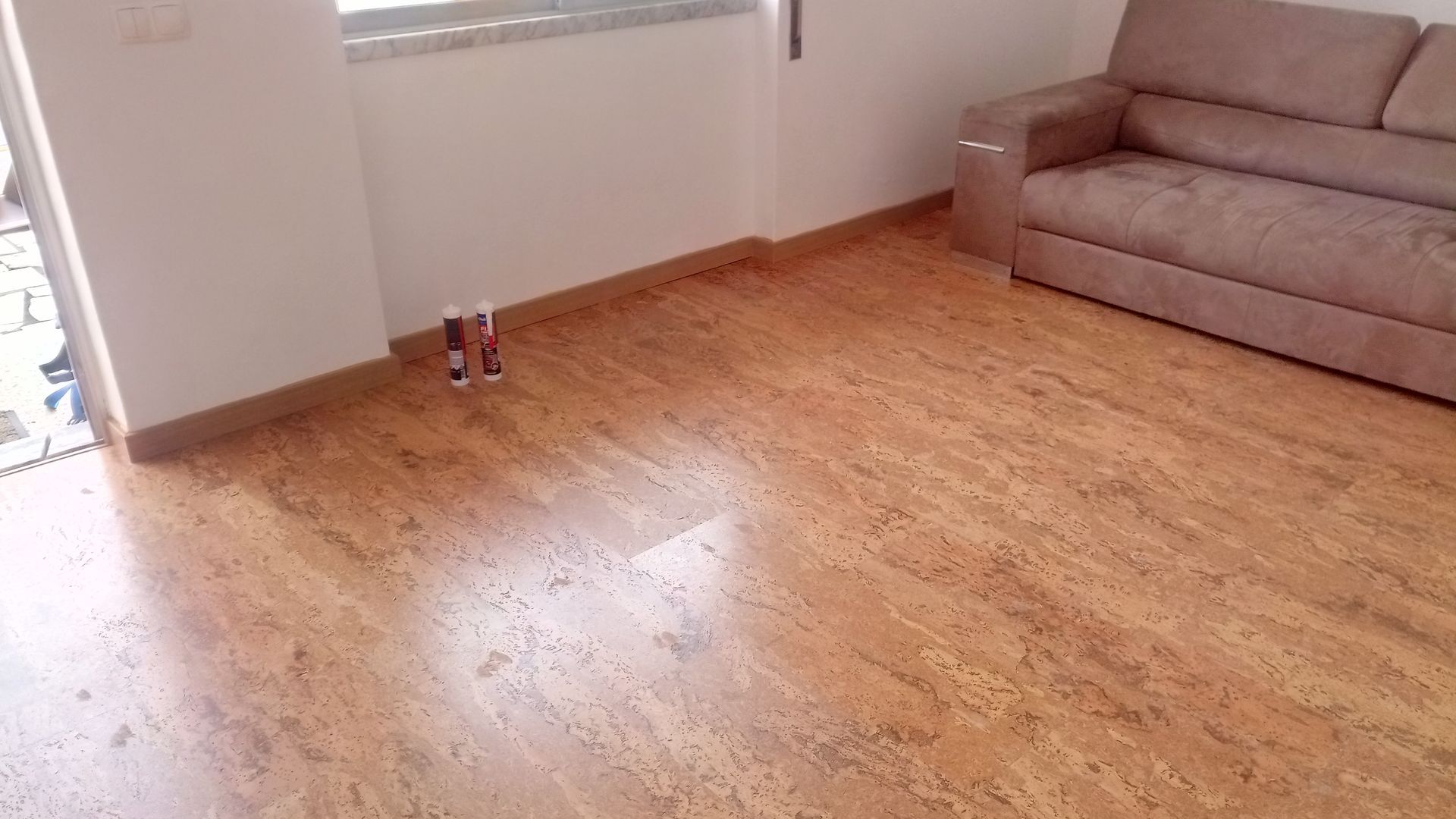 Sala com piso que imita madeira, um sofá bege e paredes brancas. Duas garrafas pequenas estão perto da porta.