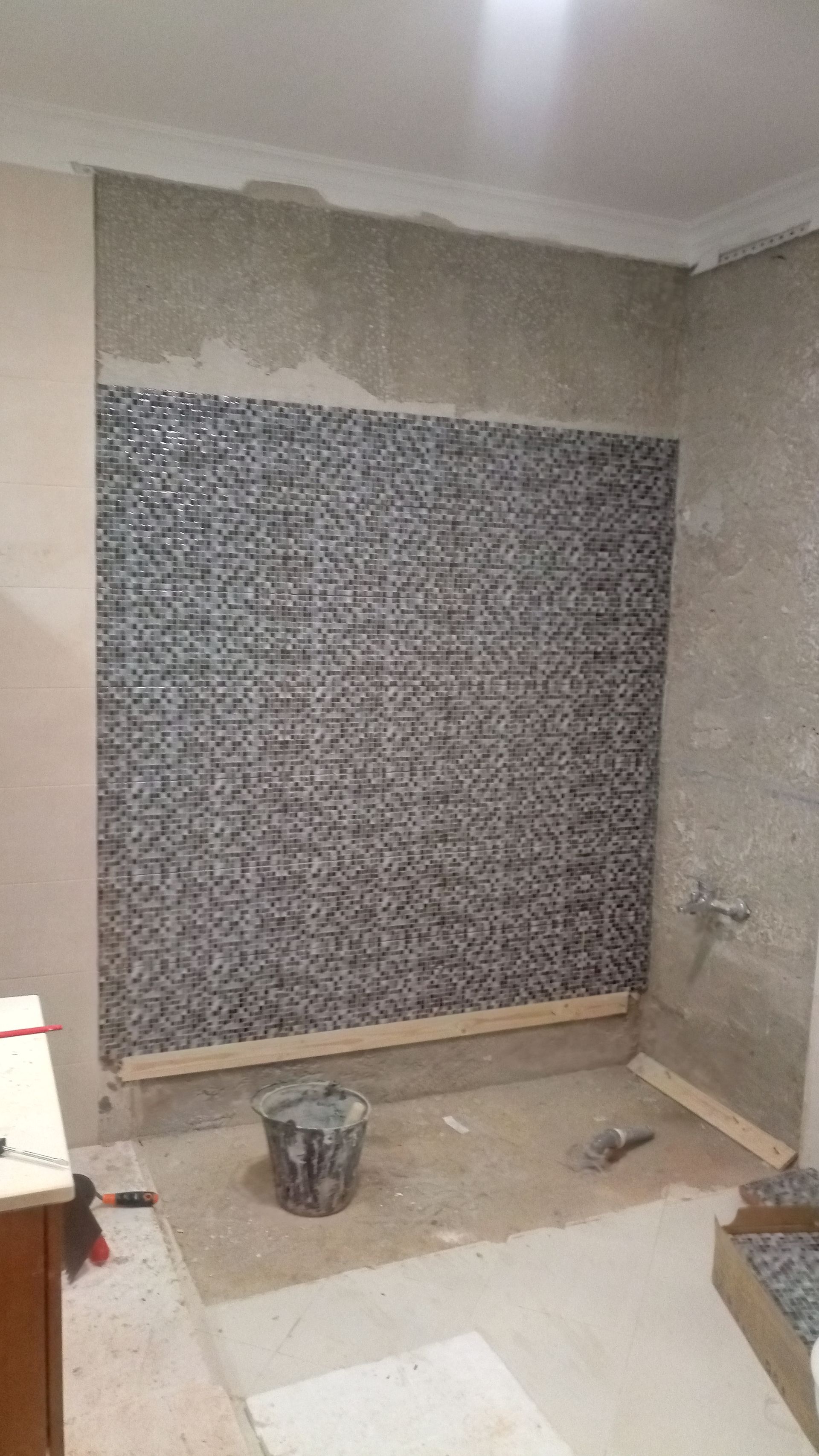 Parede do banheiro com azulejos de mosaico parcialmente instalados; obra em andamento.