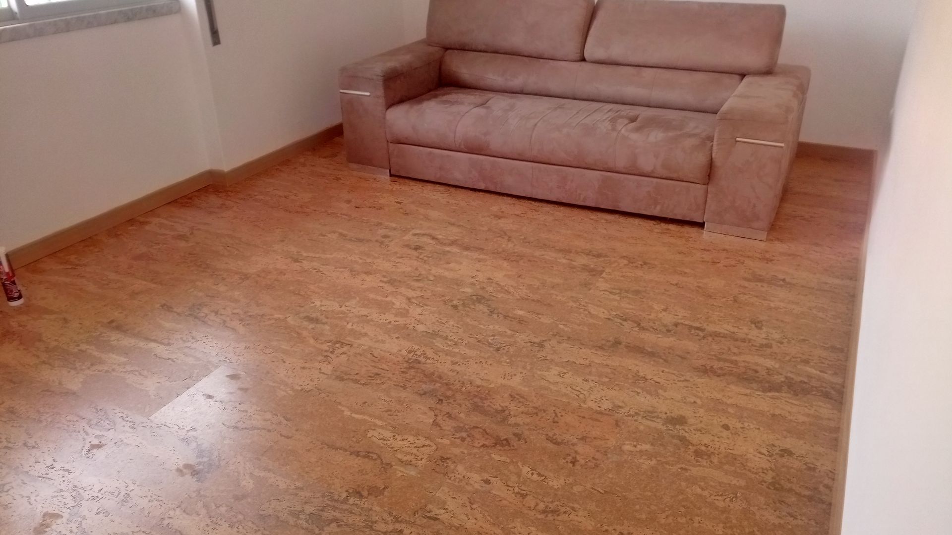 Quarto com piso de cortiça, um sofá bege encostado na parede e uma janela.