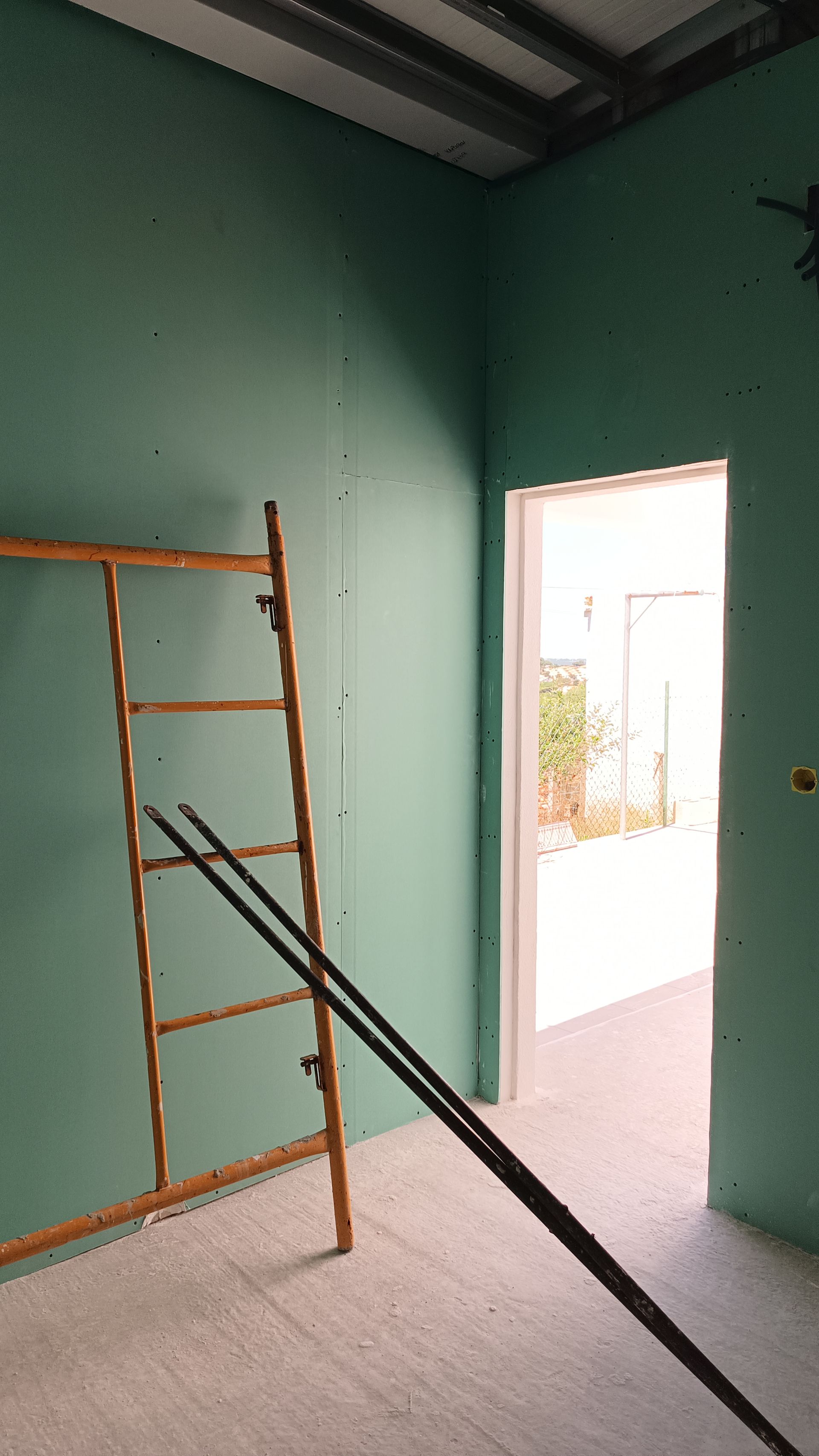 Um cômodo em construção com paredes de gesso verde, uma porta aberta e uma escada de madeira encostada na parede.
