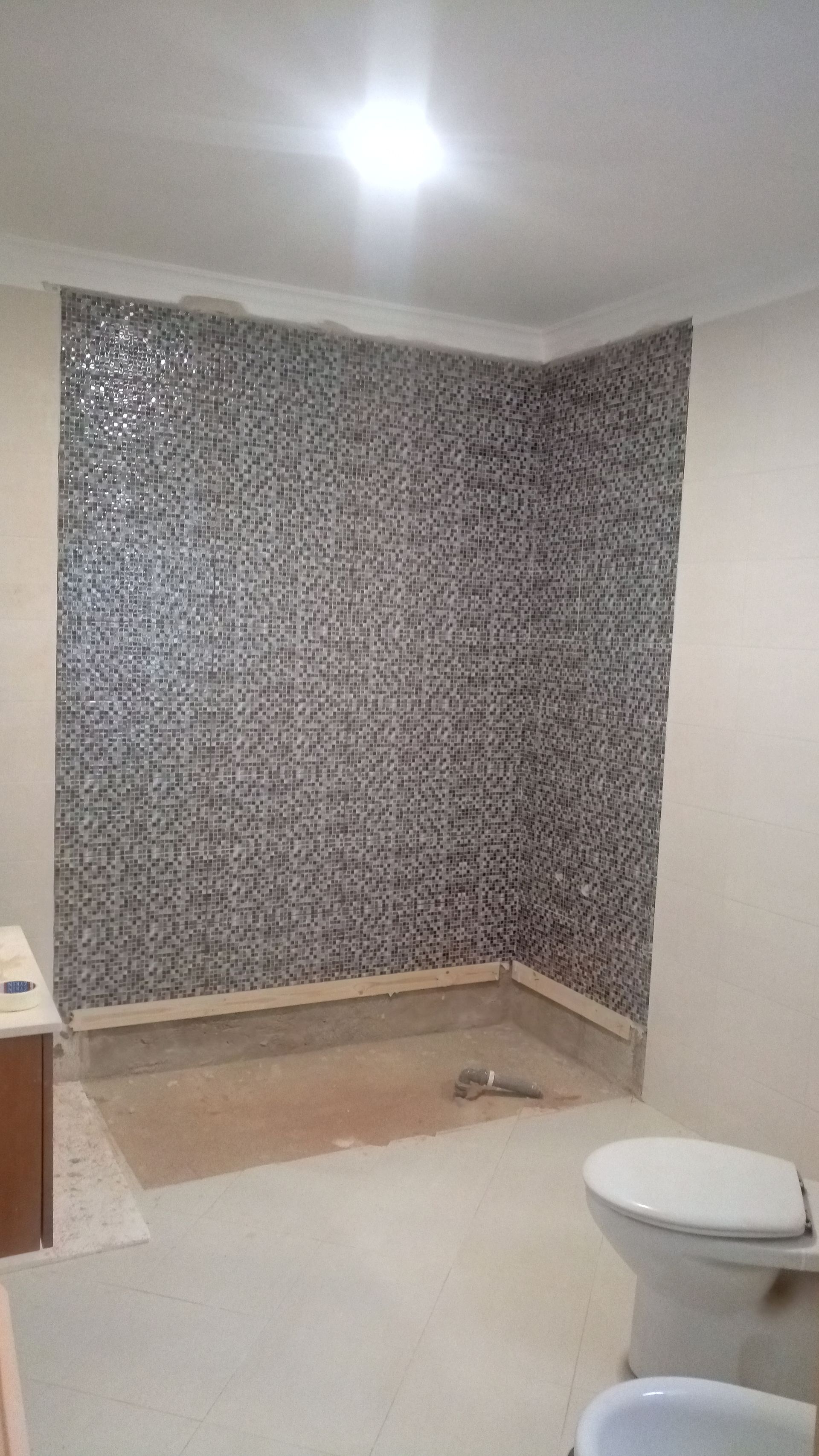 Banheiro com parede do chuveiro revestida com azulejos em mosaico, piso inacabado, vaso sanitário e pia.