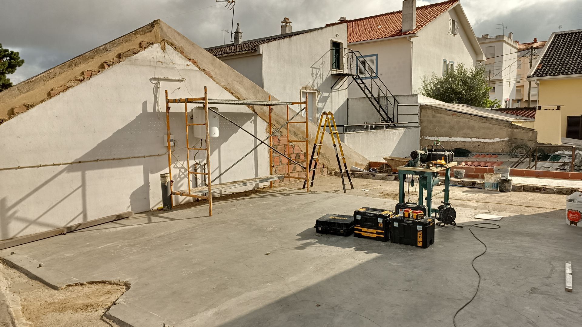 Canteiro de obras com andaimes e equipamentos. Prédios brancos e um pátio de concreto.