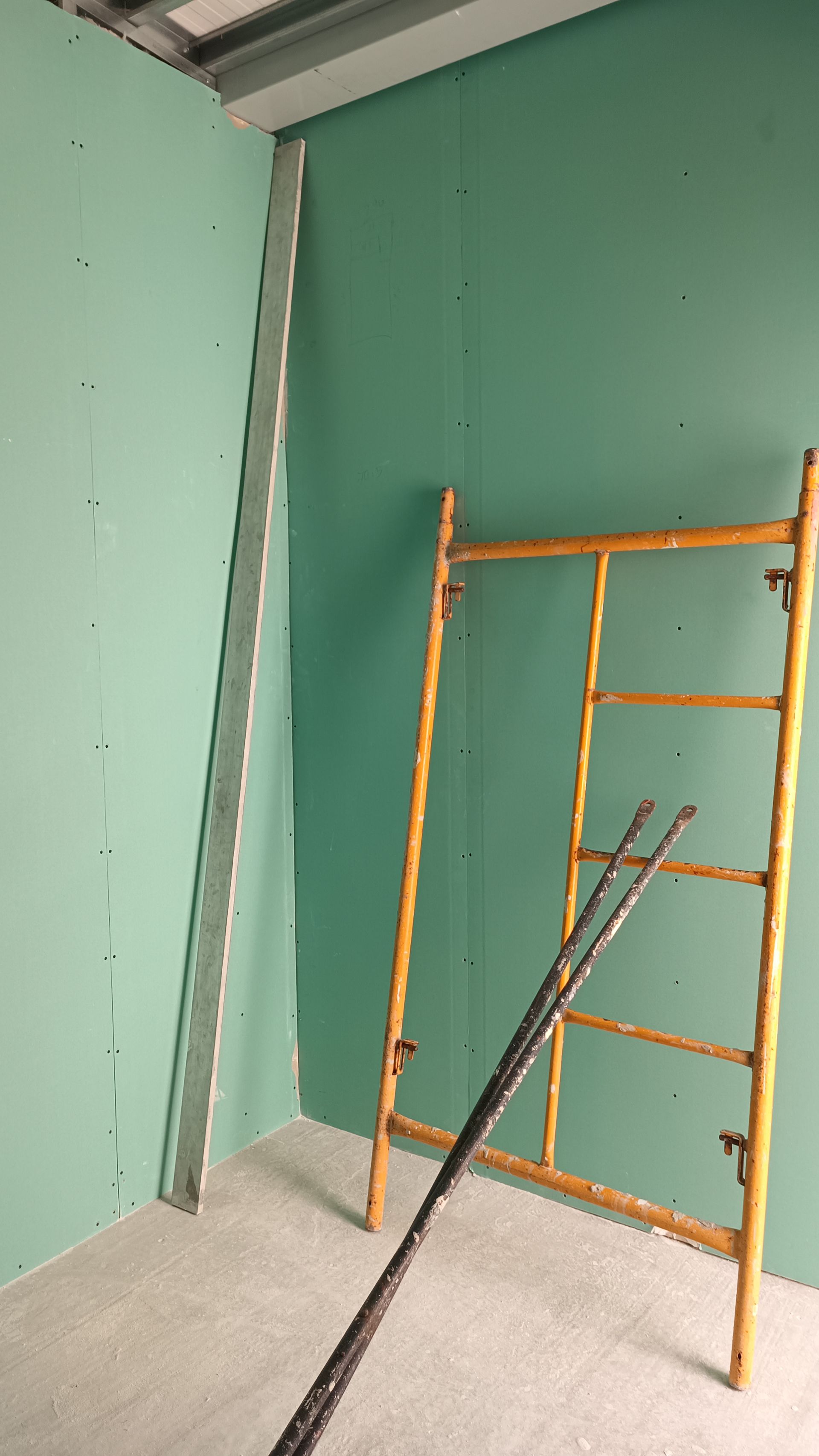 Parede de gesso verde em uma sala em construção, com um andaime amarelo e uma régua de metal.