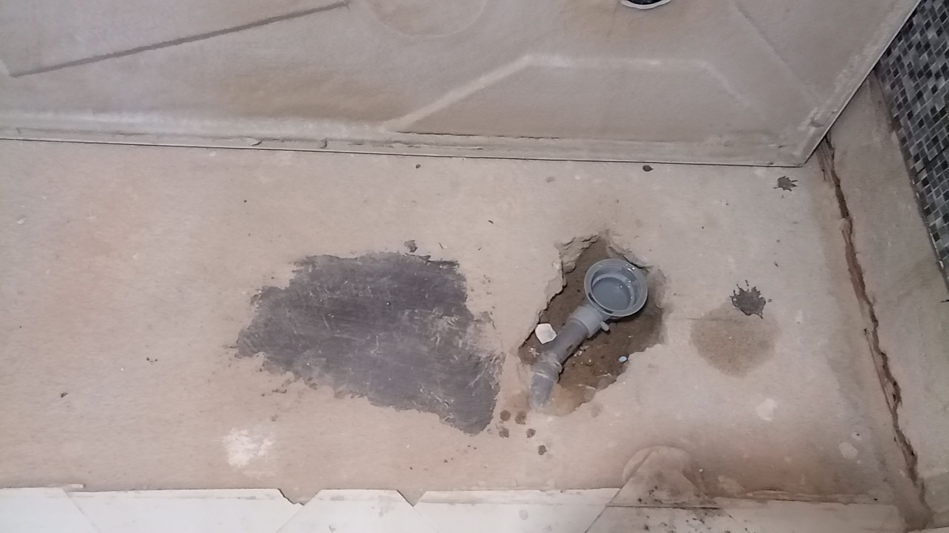 Piso do chuveiro danificado com ralo, apresentando manchas de água e remendo.