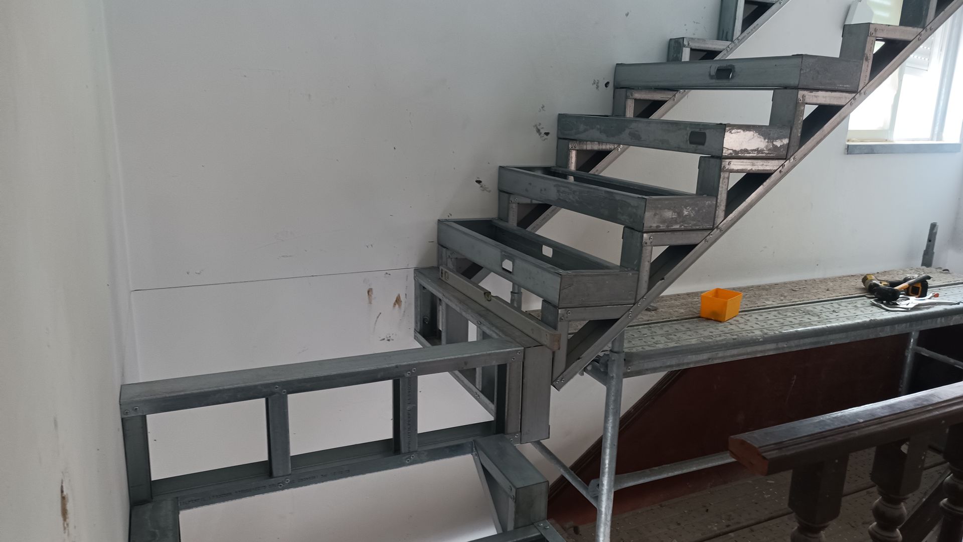 Escada metálica em construção, com espelhos abertos e estrutura de suporte, contra uma parede branca.