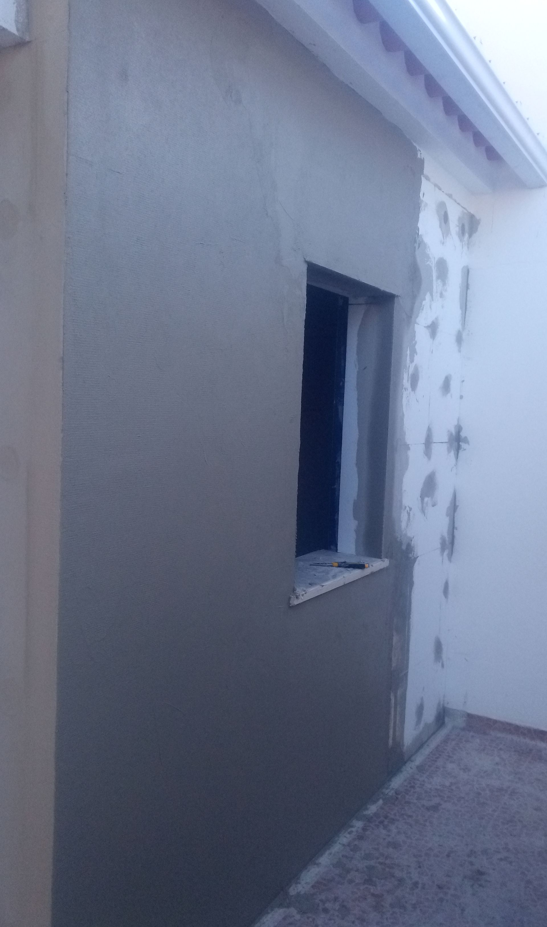 Parede exterior com janela, parcialmente rebocada com cimento cinza; telhado branco visível.