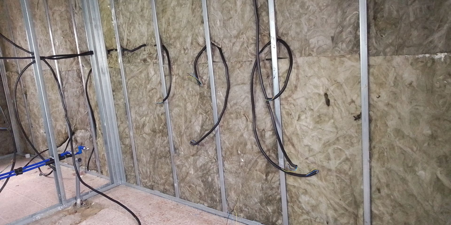 Parede interna em construção, com perfis metálicos, isolamento e fiação elétrica.