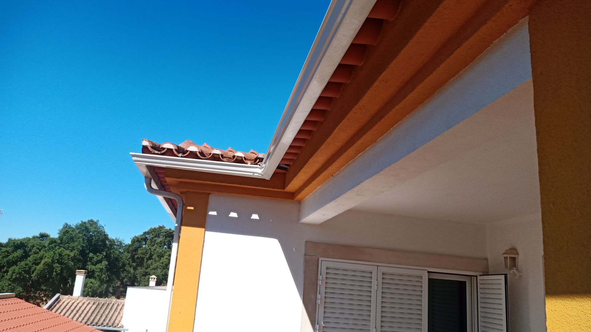 Close-up da fachada de um prédio com detalhes em laranja, telhas de terracota, parede branca e céu azul claro.