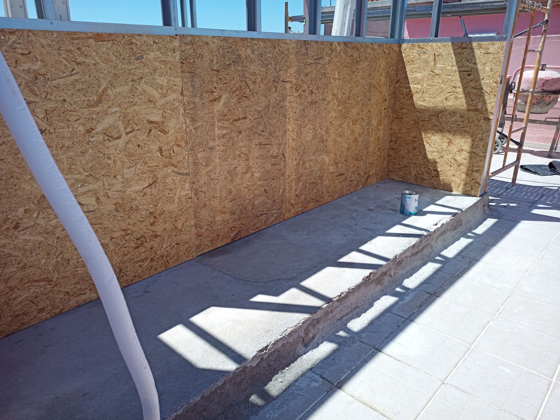 Painéis OSB instalados em um canteiro de obras. Fundação de concreto e estrutura metálica visíveis. Ambiente externo ensolarado.