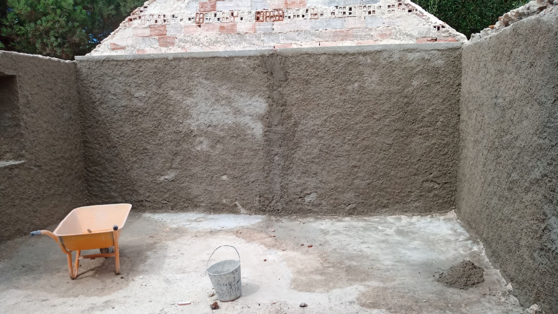 Interior de uma sala em construção, com paredes de estuque e piso de concreto. Um carrinho de mão e um balde são visíveis.