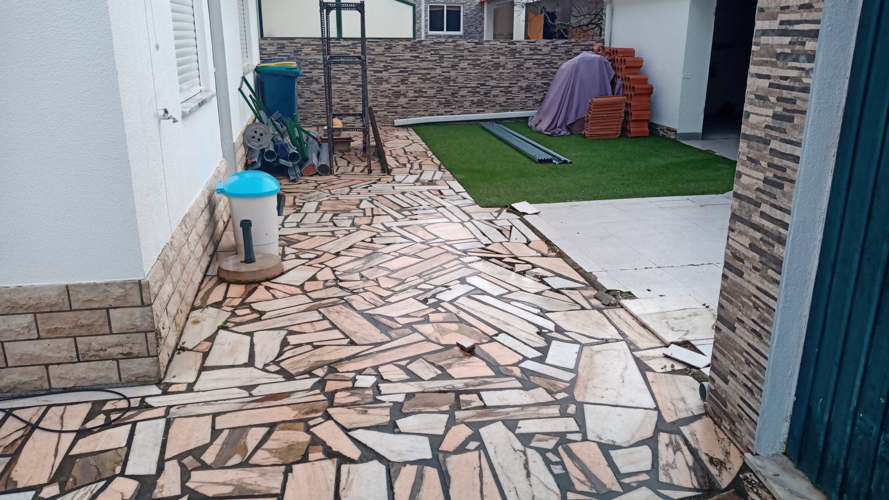 Caminho com pavimentação irregular em pedra, que leva a uma área gramada e à entrada de um edifício com materiais de construção.