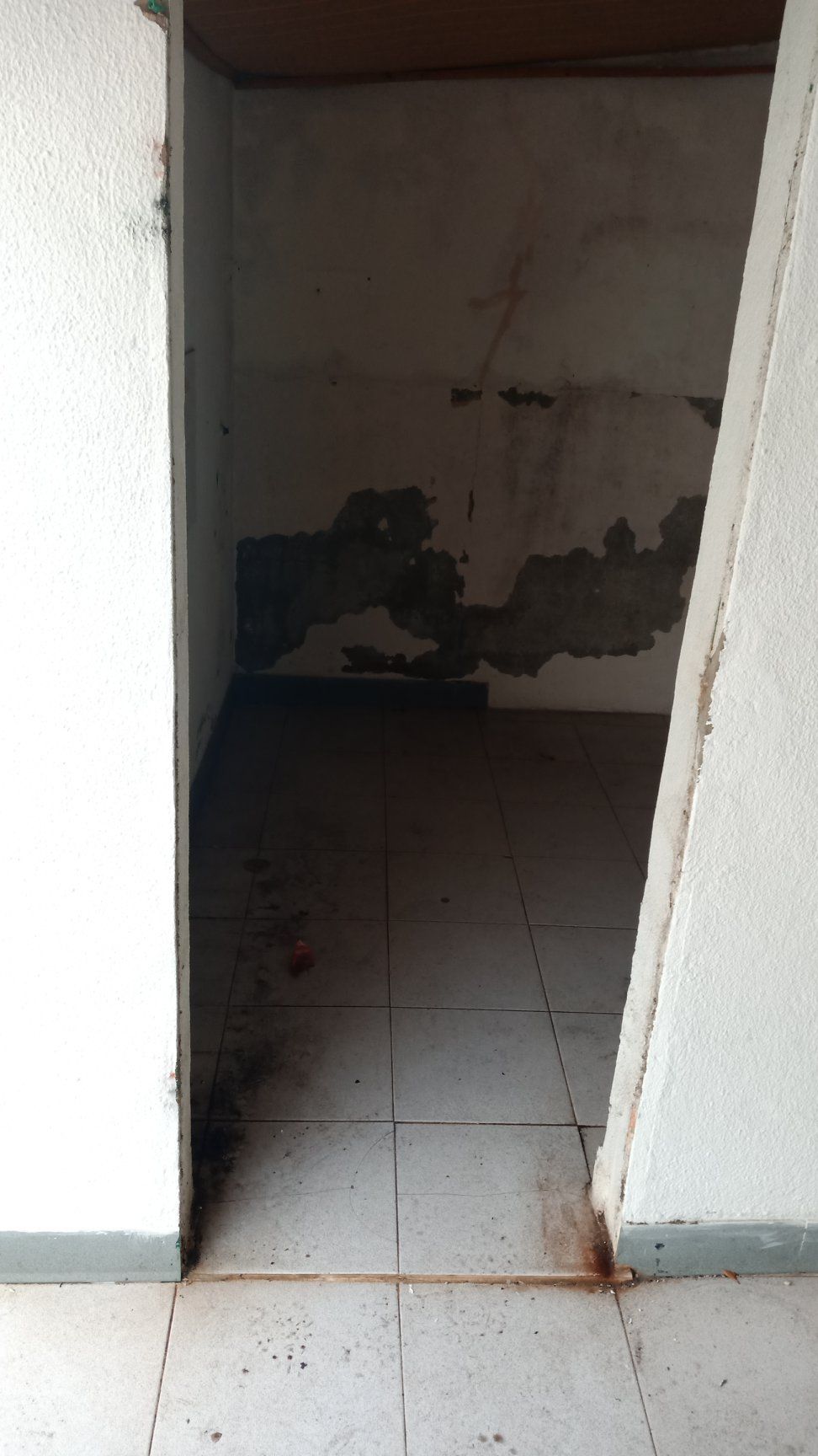 Um nicho escuro e vazio, com tinta descascando e piso de azulejos, visto através de uma porta com molduras brancas.