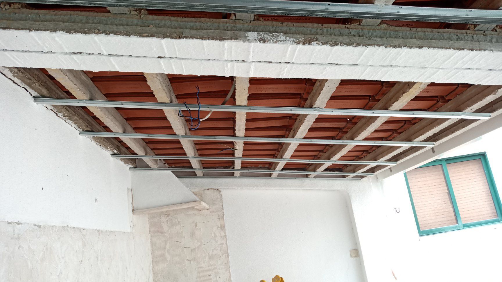 Teto em construção com telhas vermelhas e estrutura metálica. Paredes brancas e janela presentes.