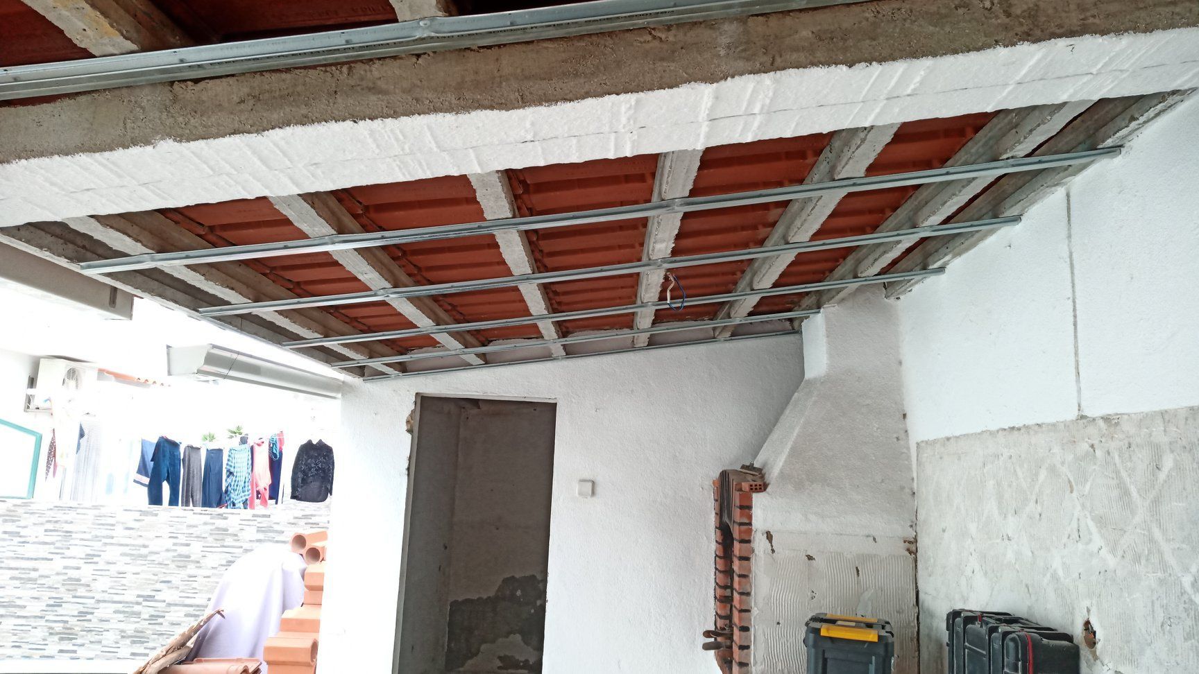 Teto em construção: estrutura metálica, telhas vermelhas, tijolo aparente, paredes brancas, ambiente externo.