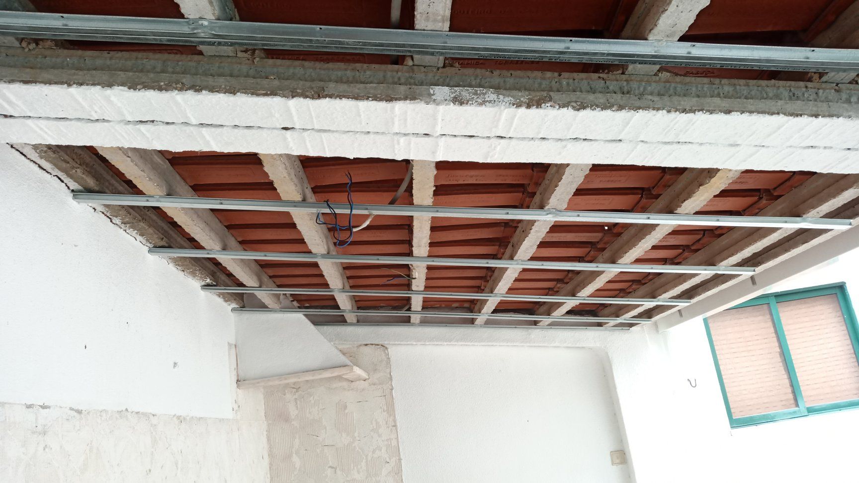Vista da construção de um teto com vigas expostas, tijolos e estrutura metálica. Também é possível ver paredes brancas e uma janela.