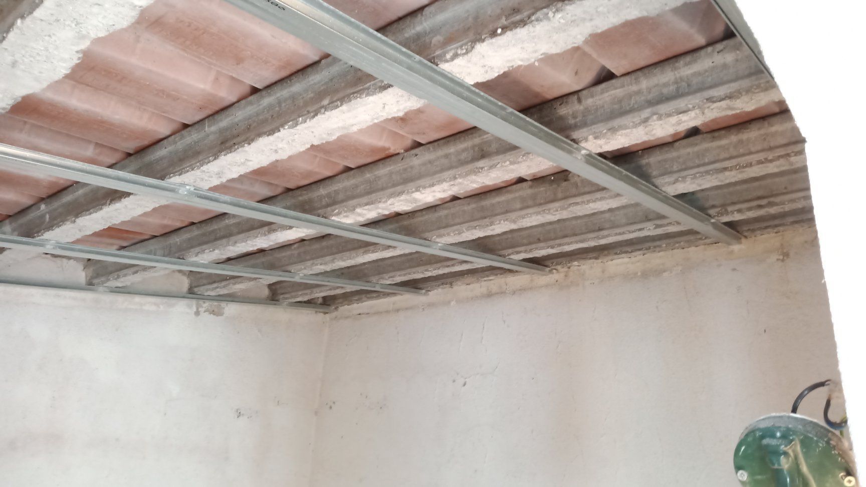 Grade metálica suspensa abaixo de um teto de azulejos vermelhos, sobre paredes de cor clara em uma sala.