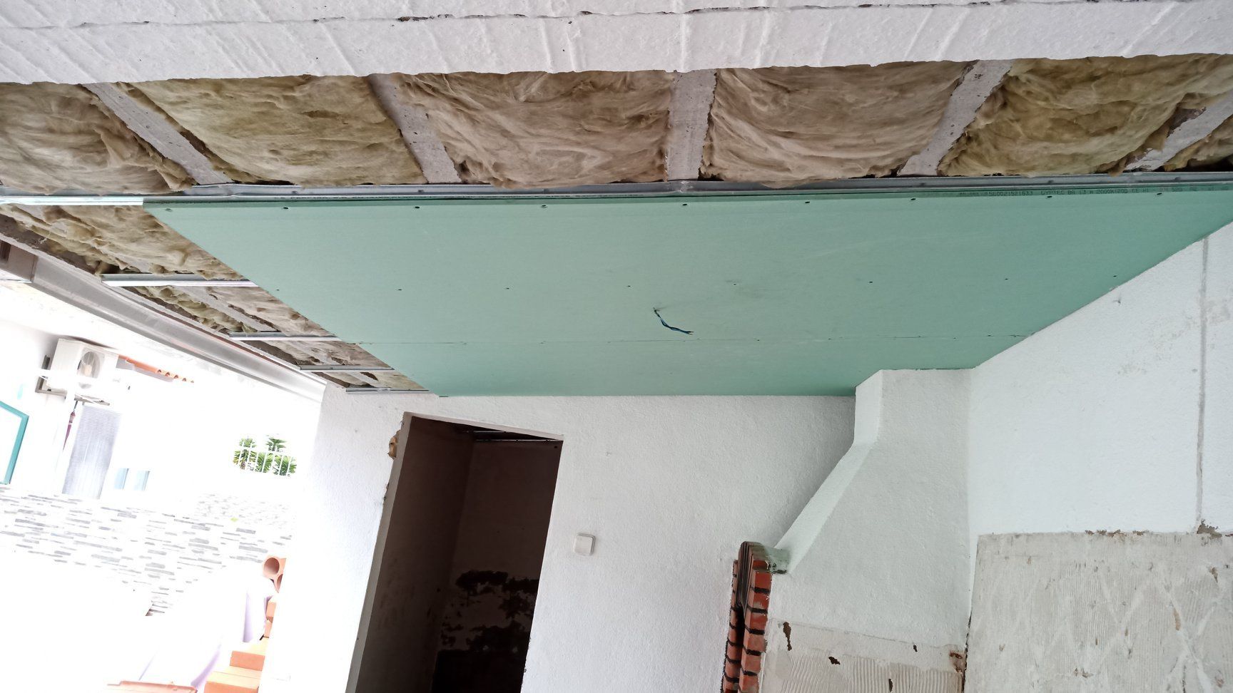 Teto em construção: placa de isolamento verde, estrutura metálica exposta e isolamento exposto acima de uma porta.