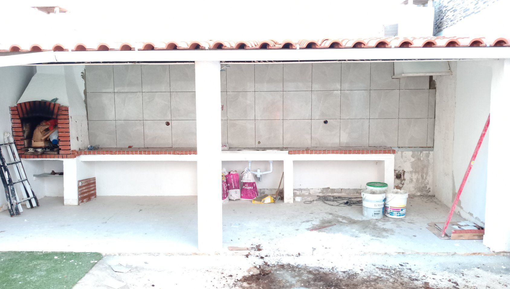 Cozinha externa em construção, com churrasqueira de tijolos, parede revestida com azulejos e bancadas. Materiais de construção visíveis.