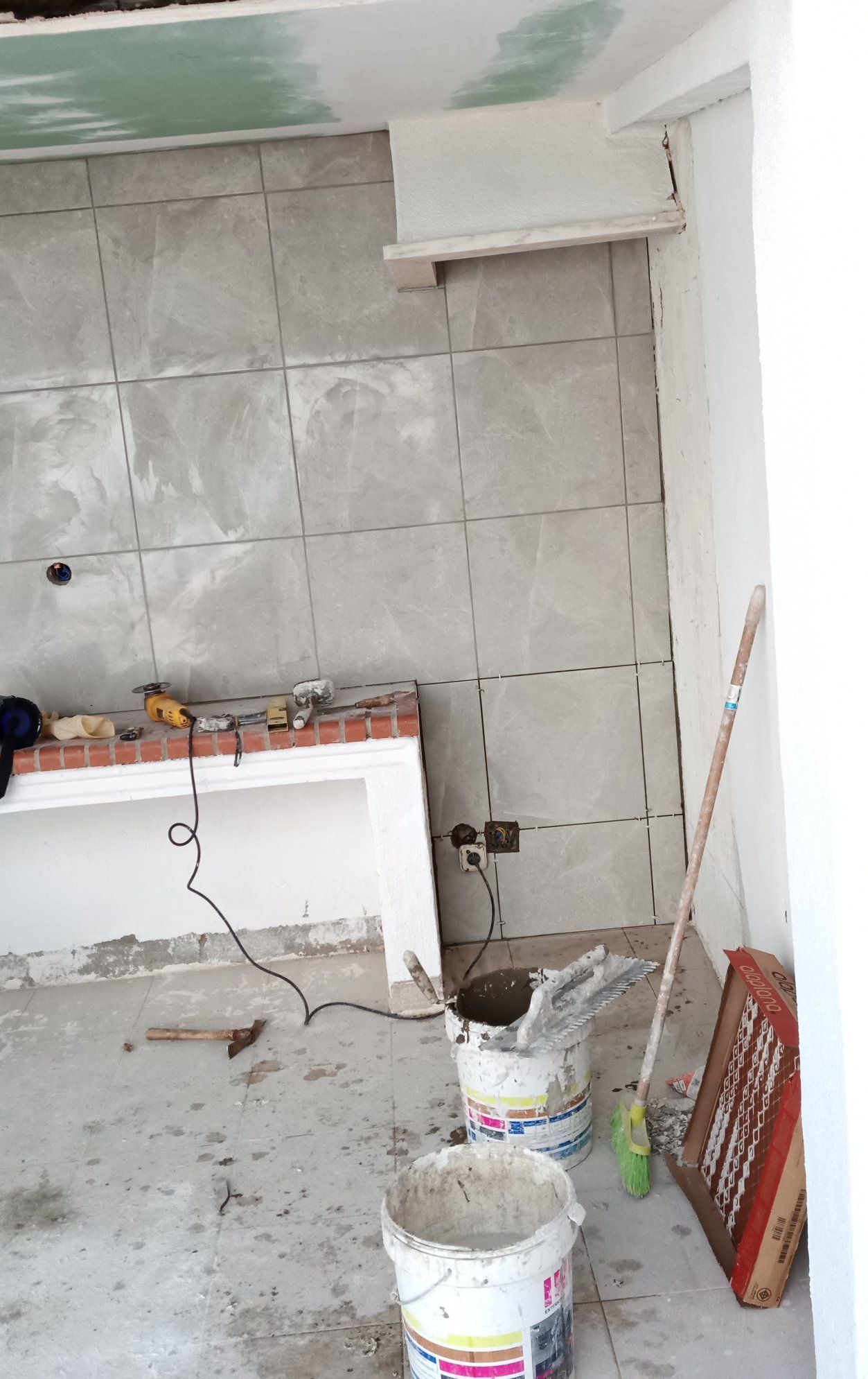 Quarto em construção com parede revestida de azulejos cinza, prateleiras brancas e baldes no chão.