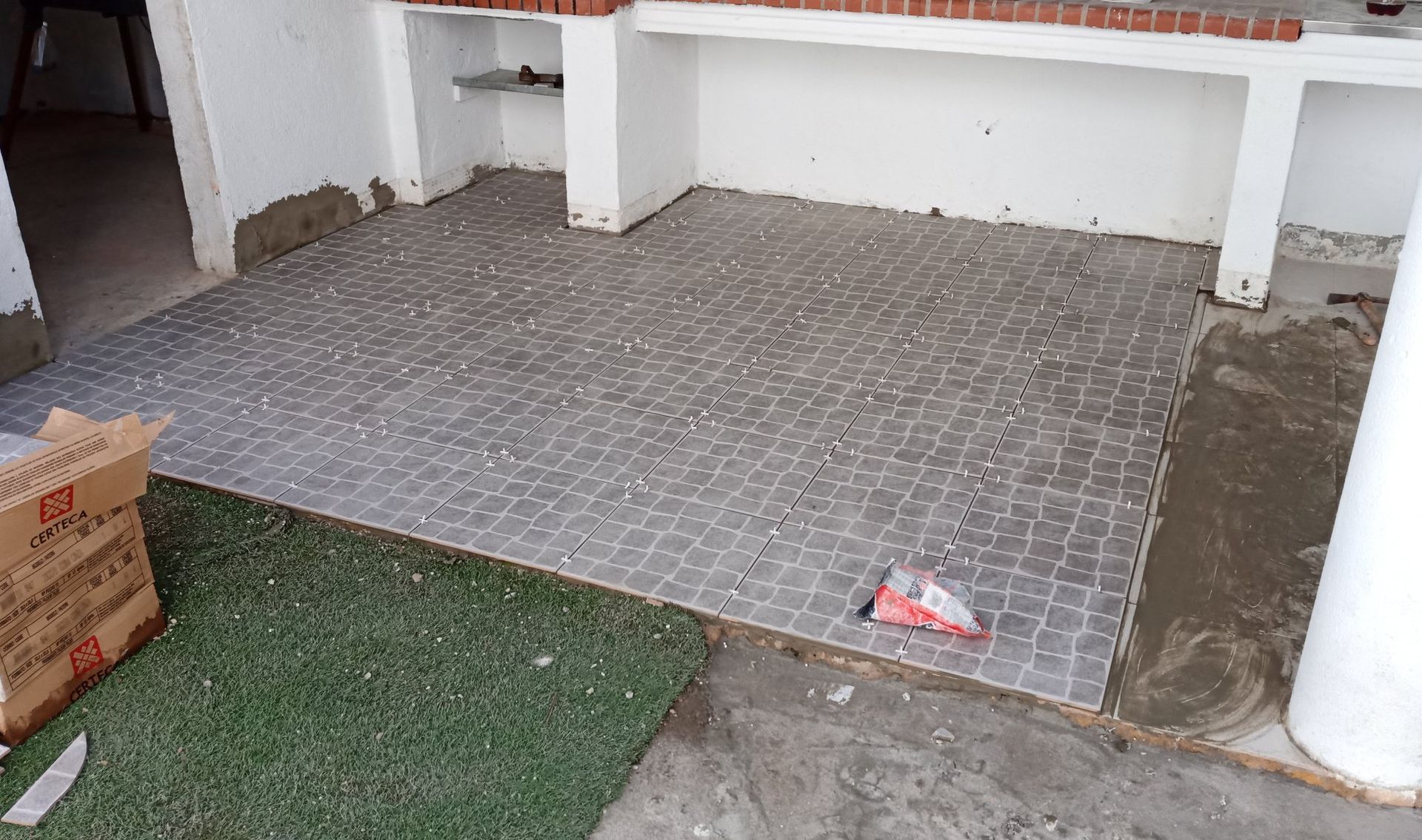 Instalação de azulejos em um pátio, com caixas e ferramentas visíveis, áreas inacabadas e um pequeno pedaço de grama.